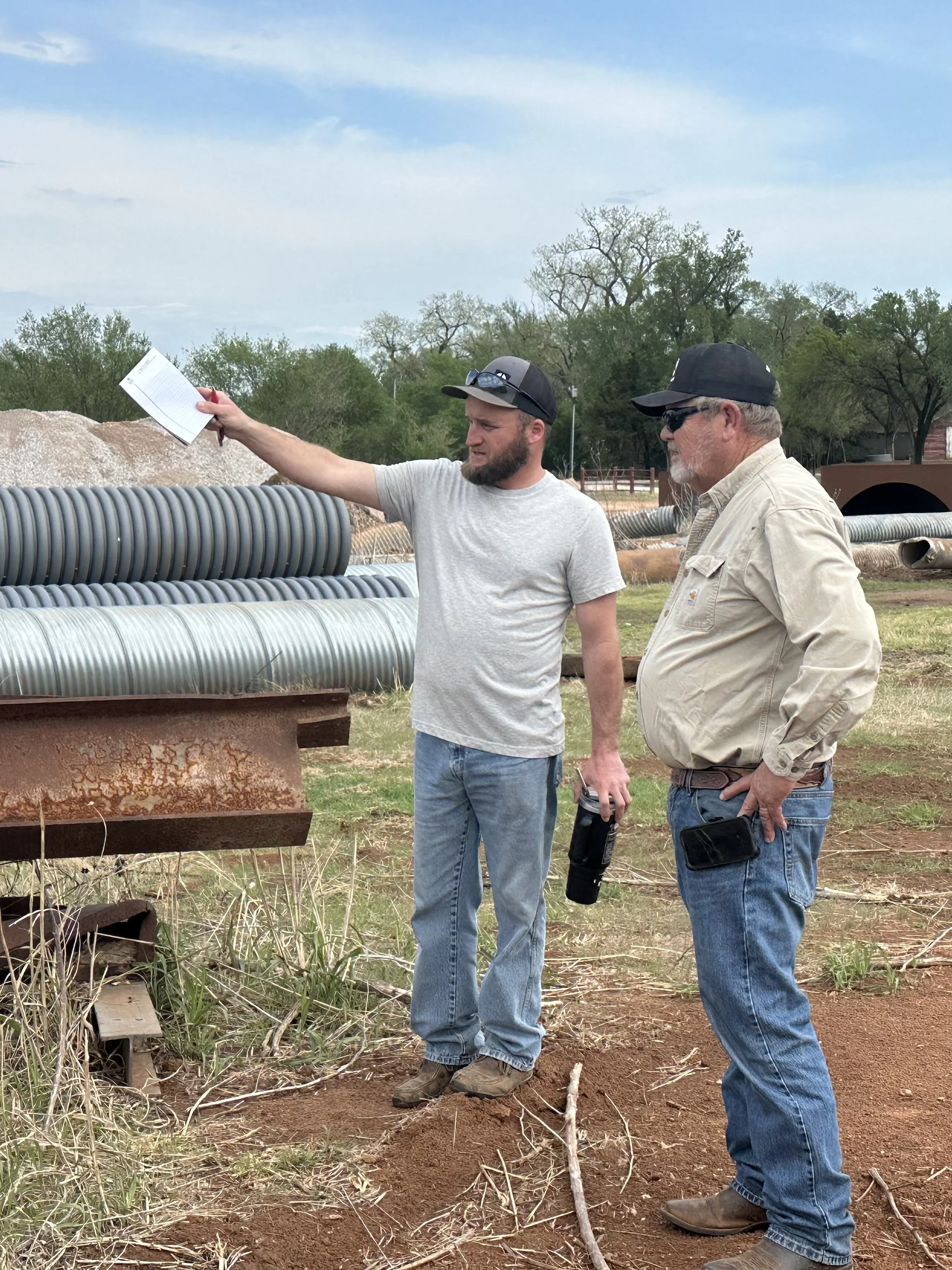 Two men standing outdoors near a large metal pipe, one holding a notebook and a drill, with trees and a partly cloudy sky in the background.