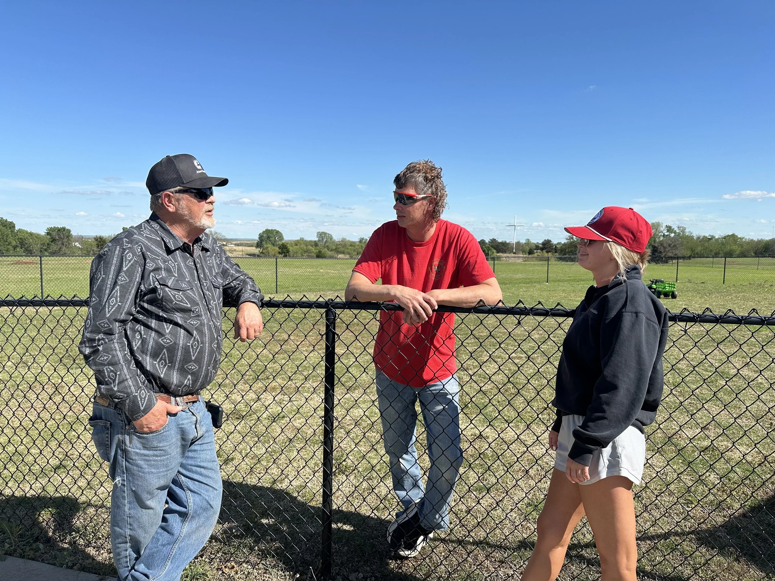 Three people having a conversation outdoors near a chain-link fence, with trees and fields in the background under a blue sky.