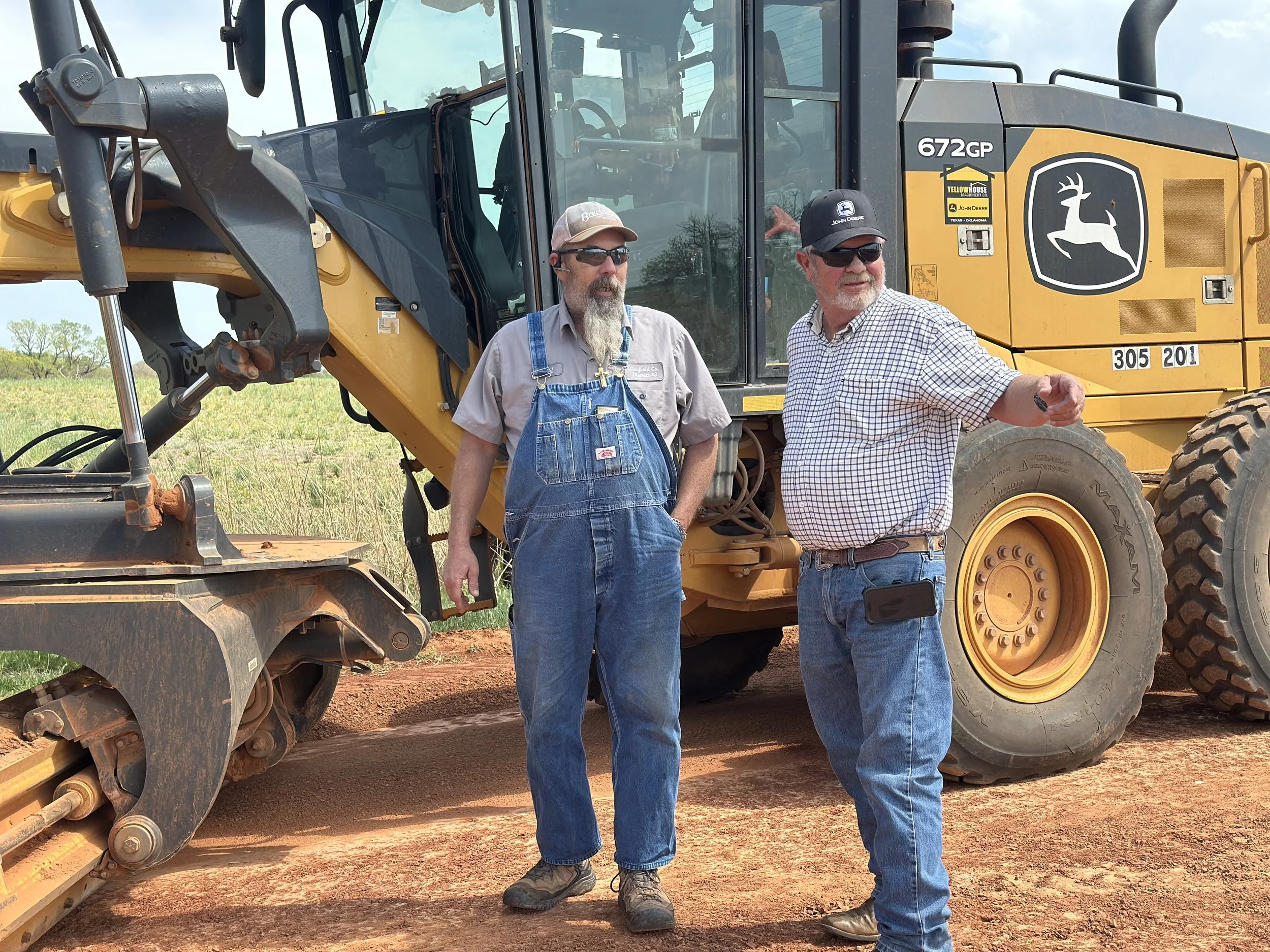Two men standing in front of a large yellow John Deere construction vehicle outdoors on a dirt road, with grassy fields and trees in the background.
