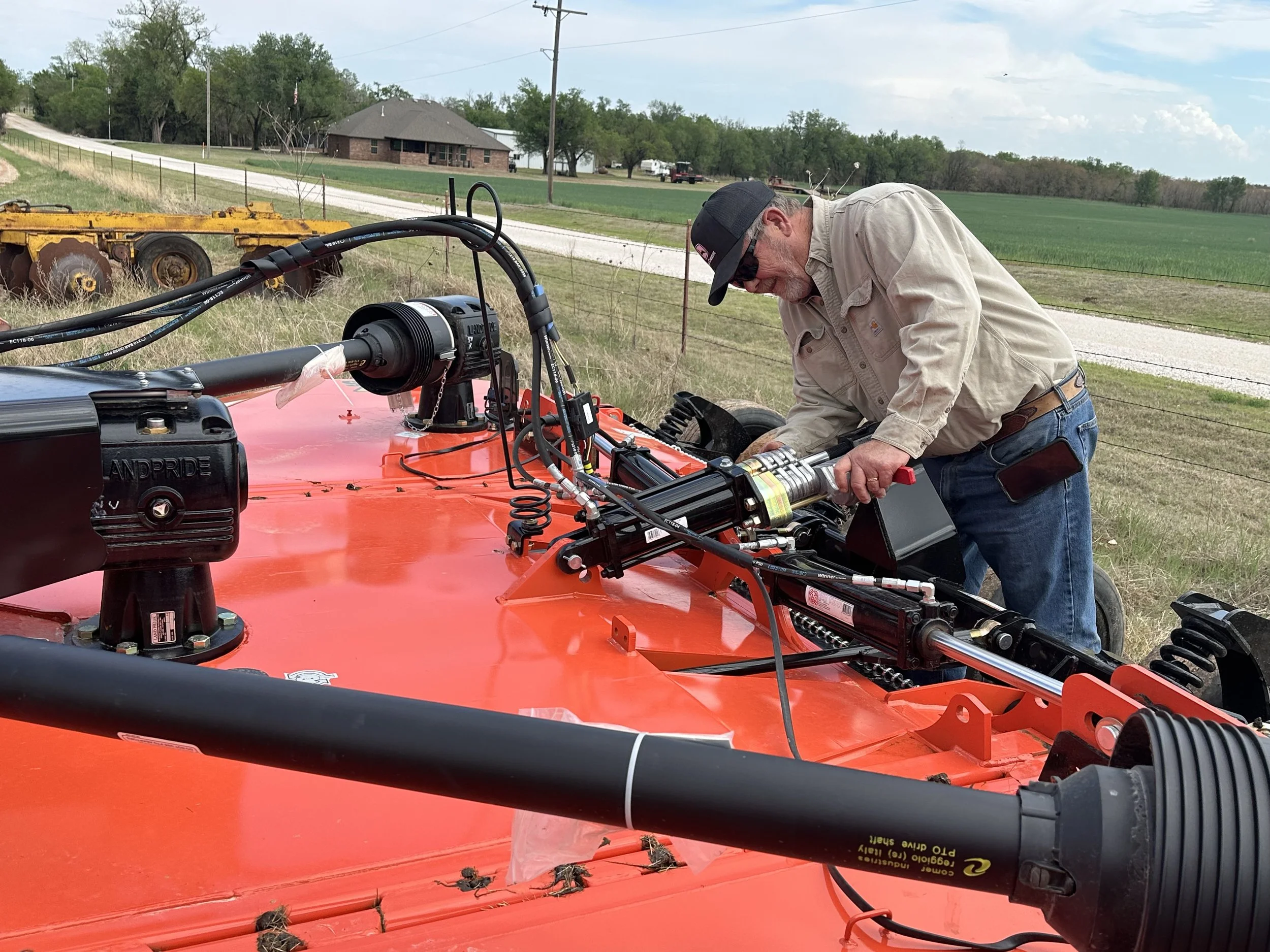 A man wearing sunglasses, a cap, and a beige shirt working outdoors on a bright red agricultural machine, with fields, a brick house, and a gravel road in the background.