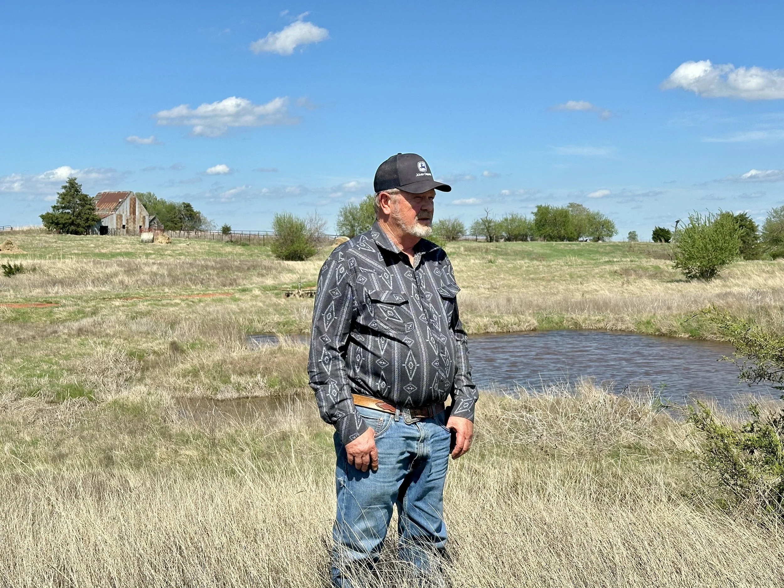 A man with a gray beard wearing a black John Deere cap, a patterned button-up shirt, and blue jeans stands outdoors near a body of water in a grassy field with trees and an old barn in the background under a partly cloudy sky.