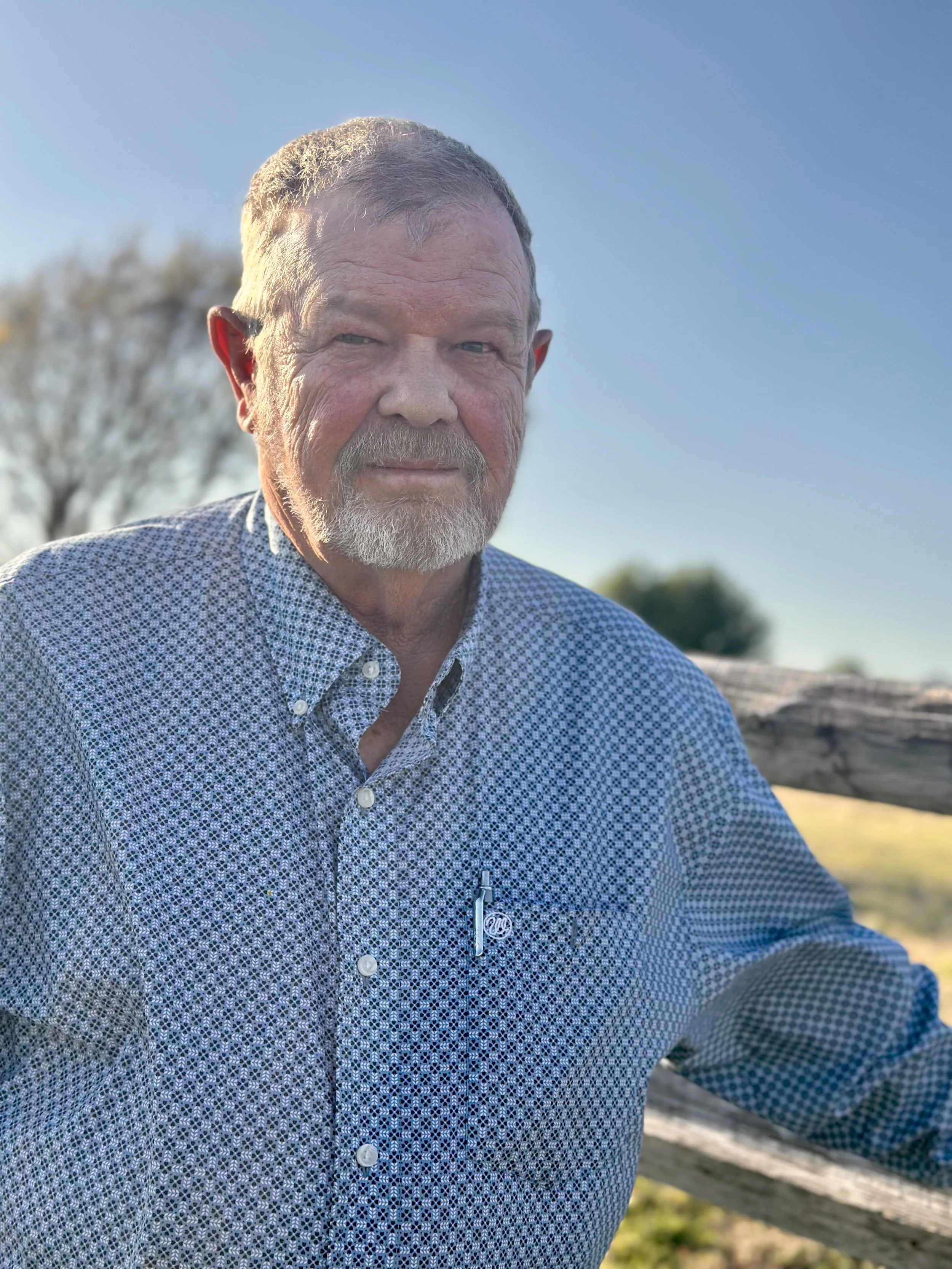 An older man with gray hair, a beard, and light skin, wearing a patterned button-up shirt, standing outdoors near a wooden fence, with a clear blue sky and trees in the background.