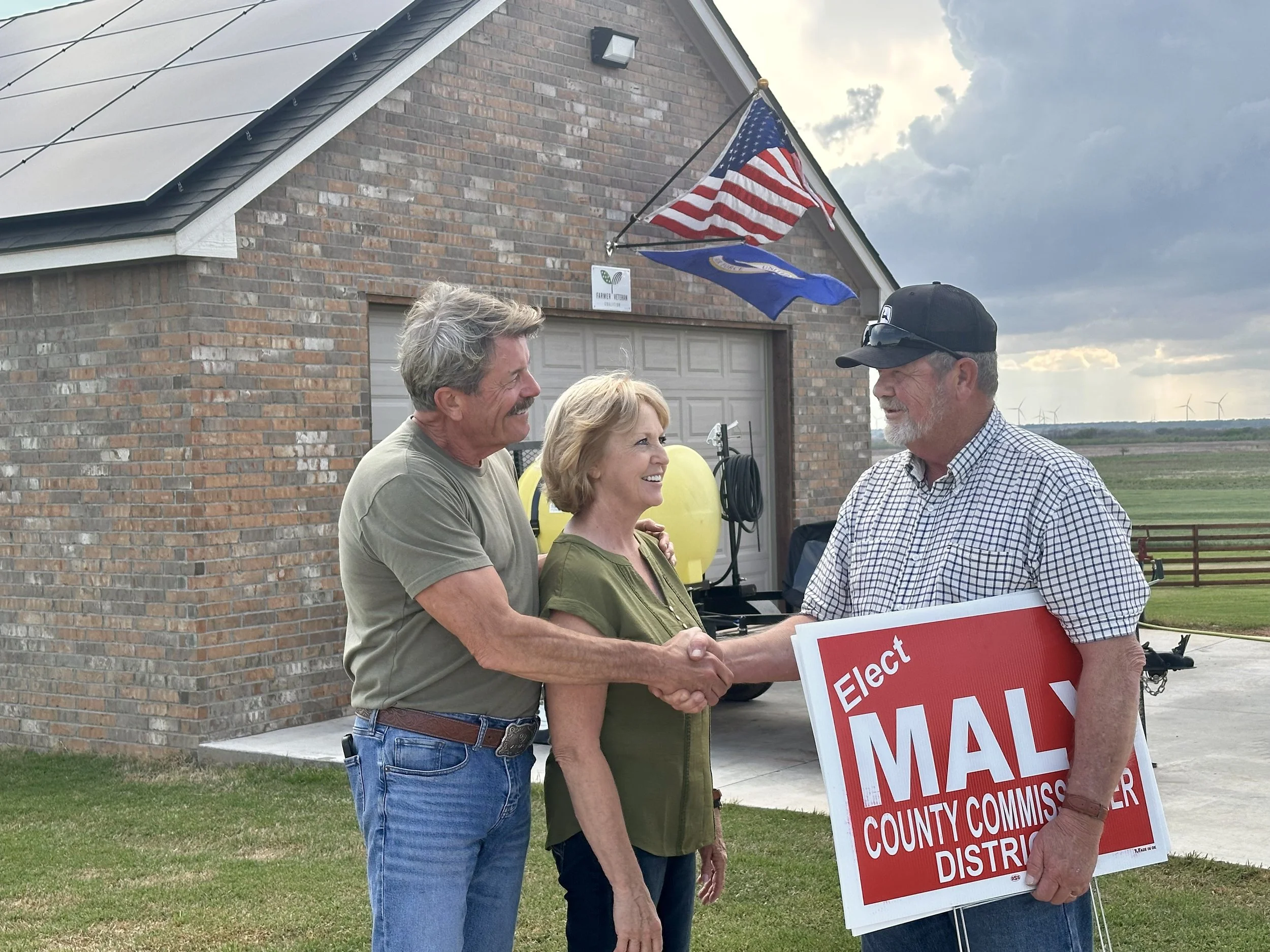 Three people outdoors in front of a brick garage, two men and one woman, shaking hands. One of the men is holding a red sign that reads 'Elect Mall' with additional smaller text. The woman is smiling and the men are engaged in conversation. There are flags mounted on the brick house, including an American flag and a state flag, and wind turbines in the distant background.