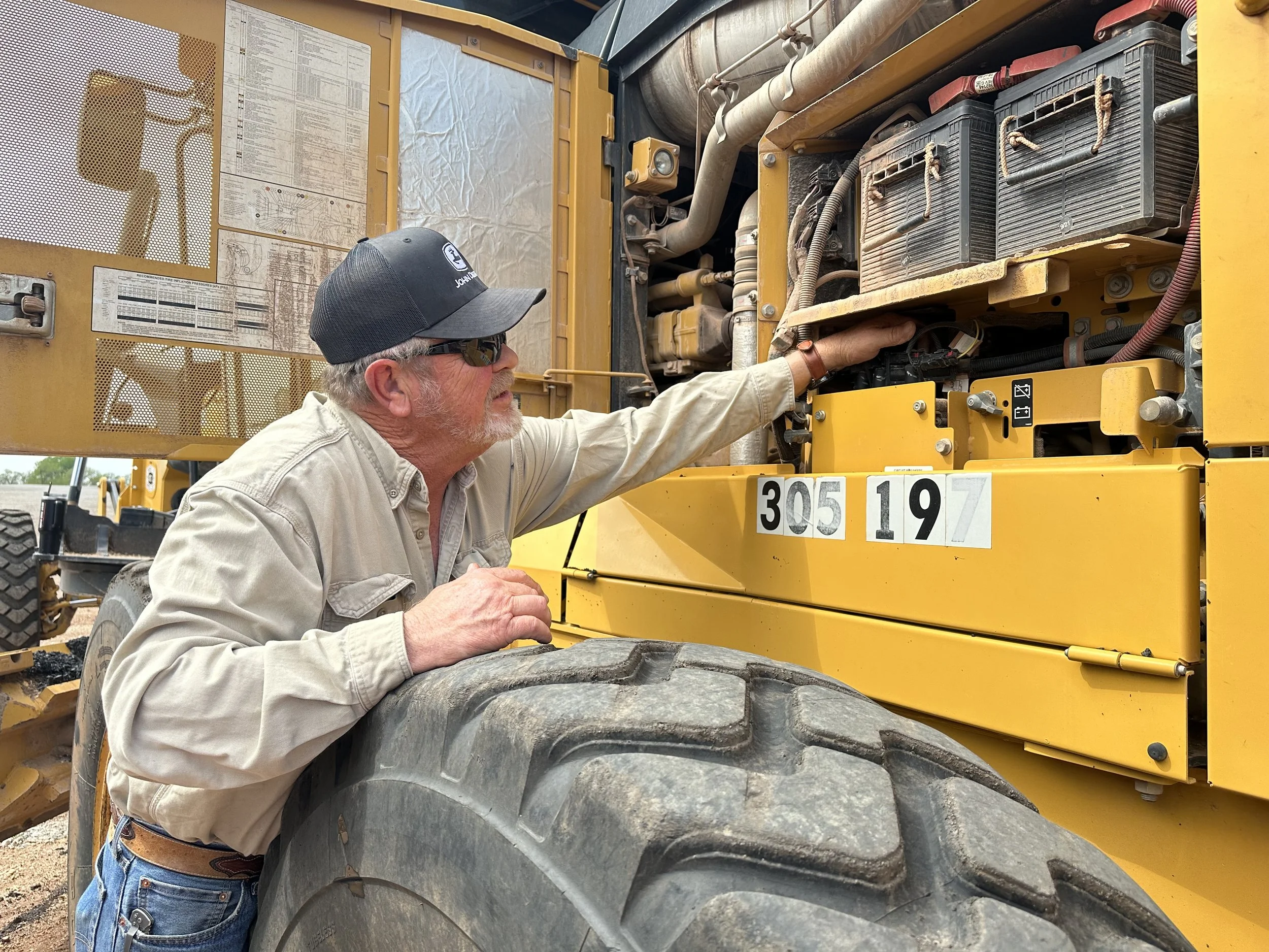 A man working on a large, yellow construction vehicle, possibly a bulldozer or loader, inspecting its engine or machinery with his hand reaching inside the engine compartment.