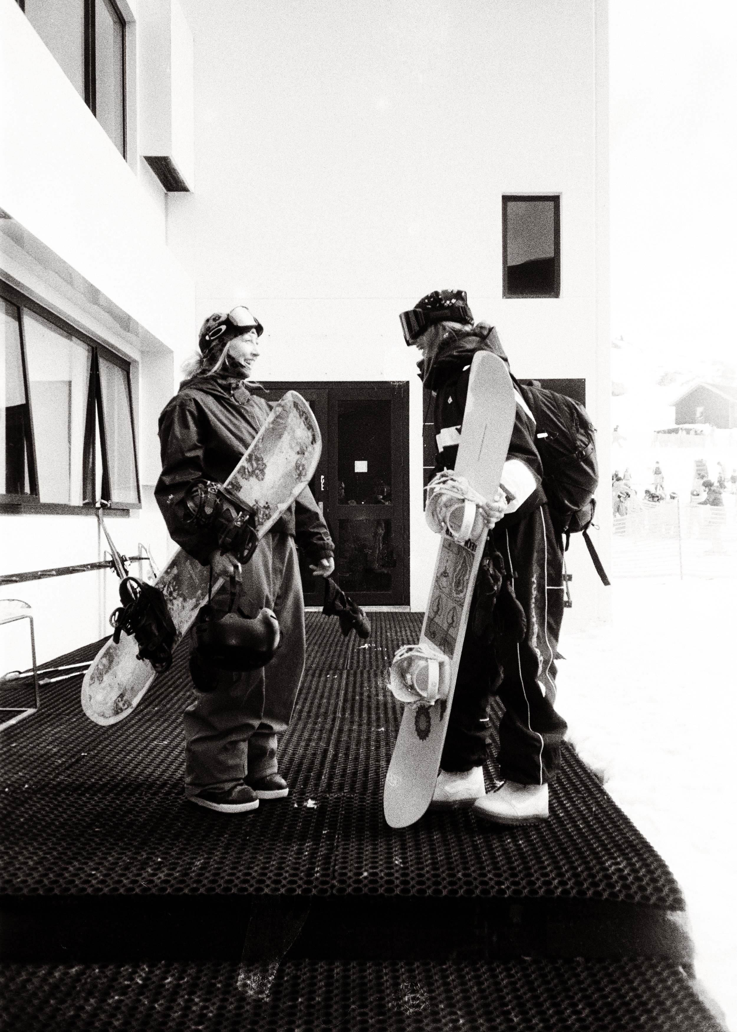 Two skiers in winter gear and helmets conversing indoors, each holding a snowboard. One is smiling. There is a window and a coat locker in the background.