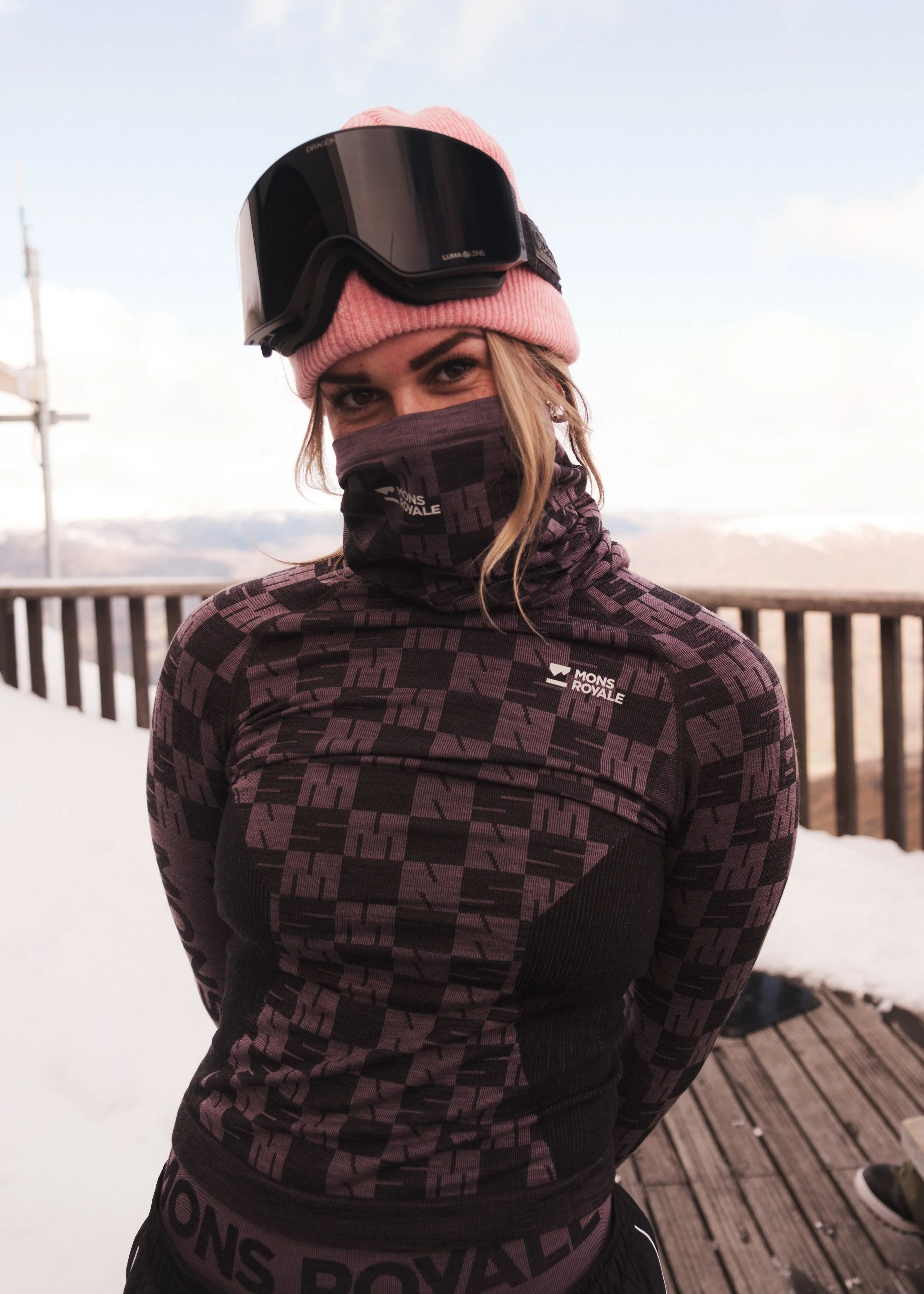 Woman wearing a pink beanie, ski goggles, and a face covering, dressed in a printed long-sleeve shirt outdoors near snow and a wooden deck with mountains in the background.