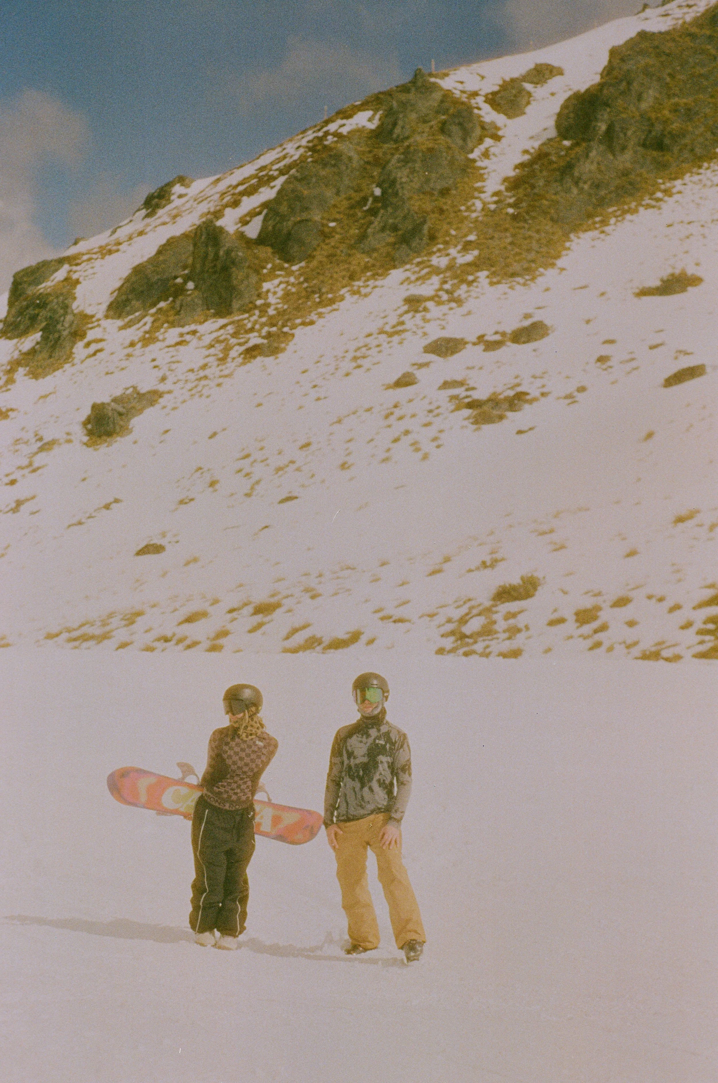 Two people in snow-covered landscape with a mountain in the background, one holding a snowboard and the other wearing sunglasses and a patterned hoodie.