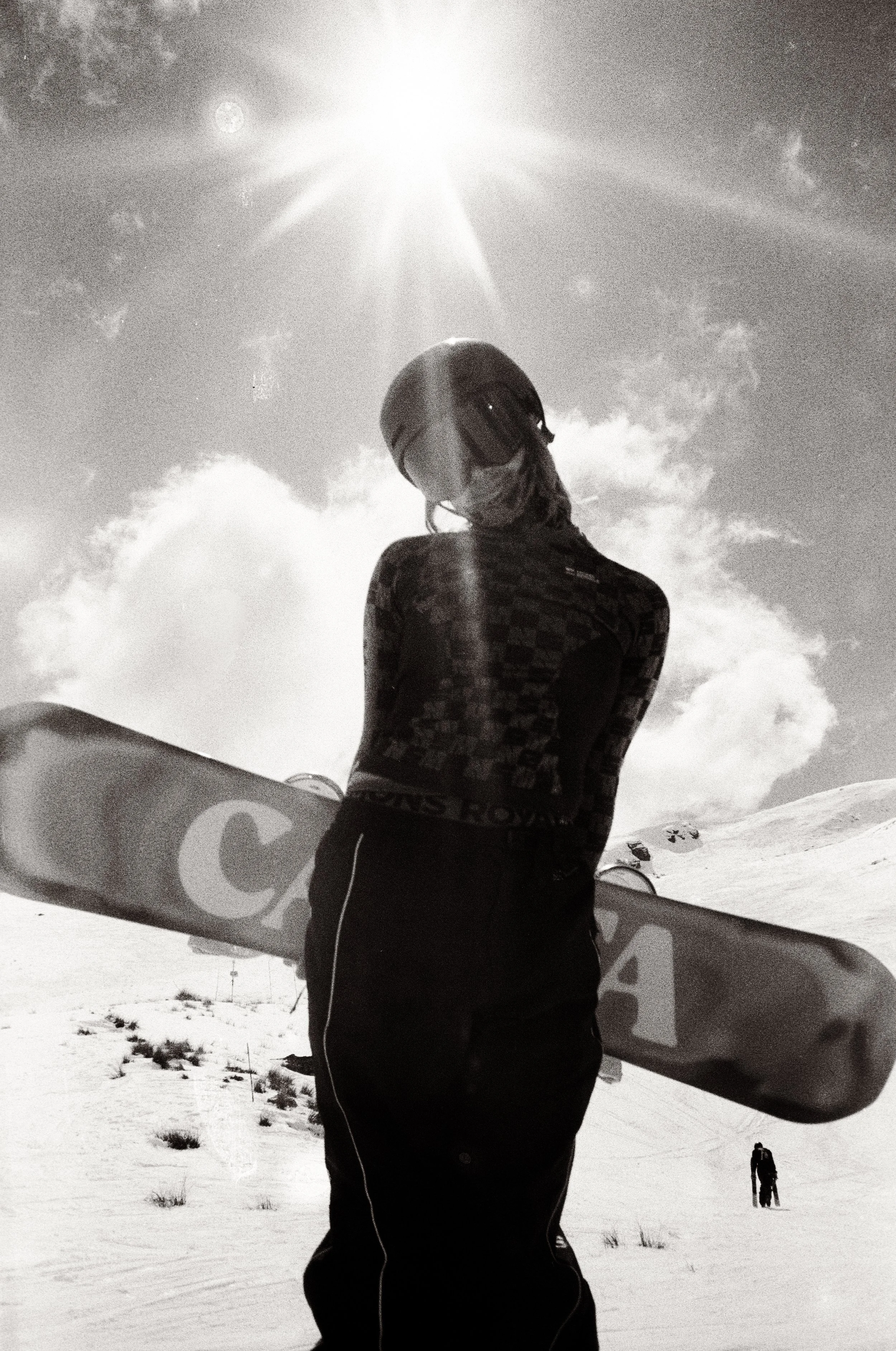 A person in the snow wearing a helmet, sunglasses, and a mask, holding a snowboard, with snow-covered ground and another person in the distance, on a bright, partly cloudy day.