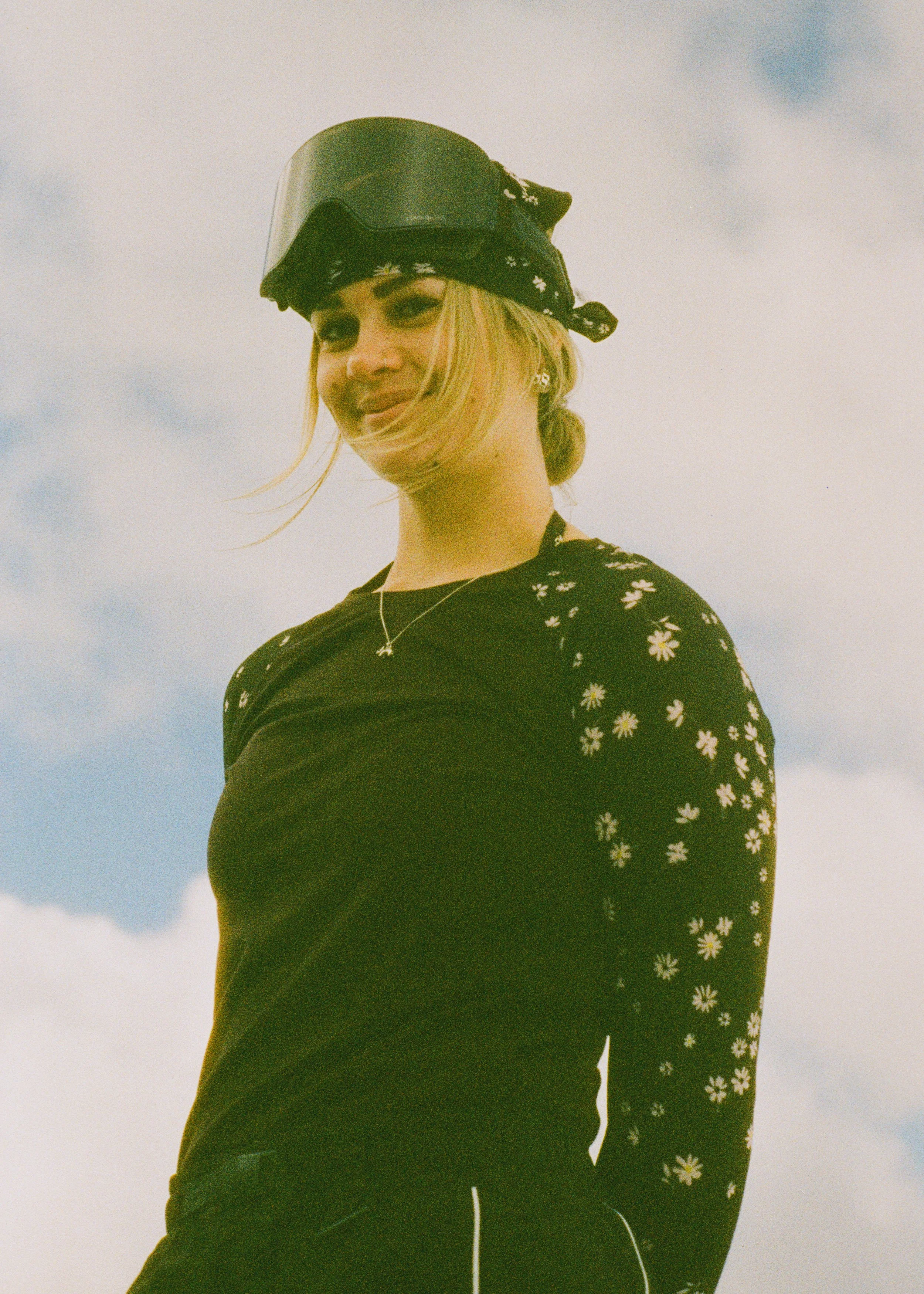 A woman with blonde hair wearing a black shirt with a white daisy pattern, a silver star necklace, black sunglasses, and a helmet with goggles, stands outdoors against a sky with clouds.