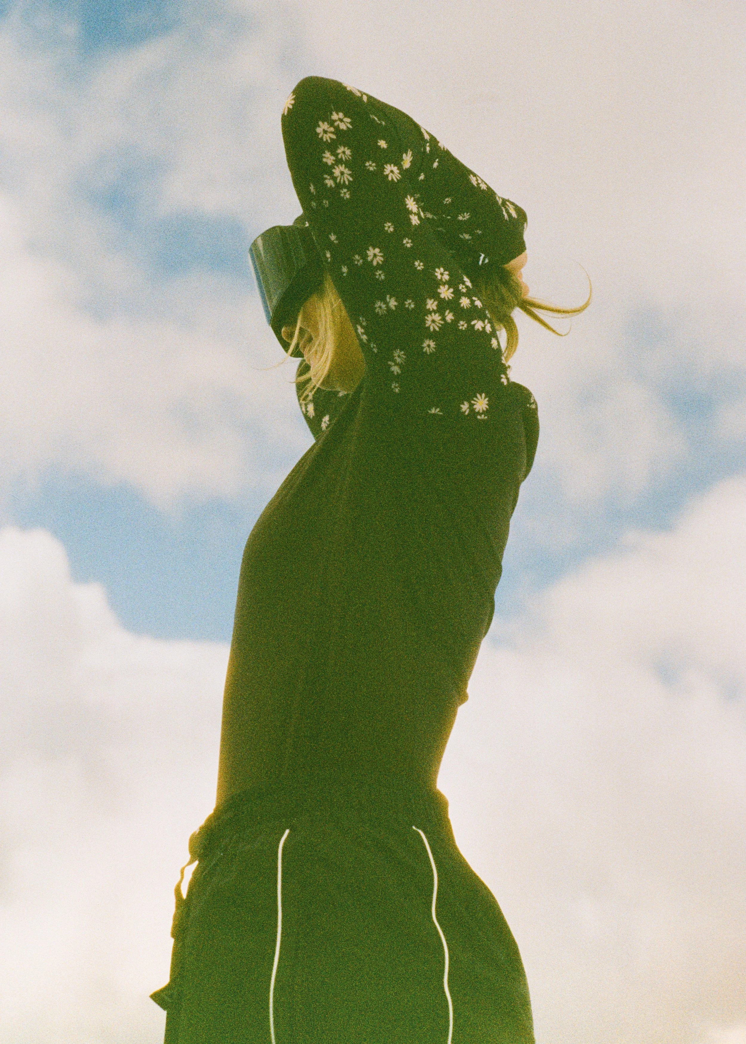 A person with long blonde hair wearing a black hoodie with white daisies, black shorts with white piping, and headphones, standing outdoors against a partly cloudy sky.