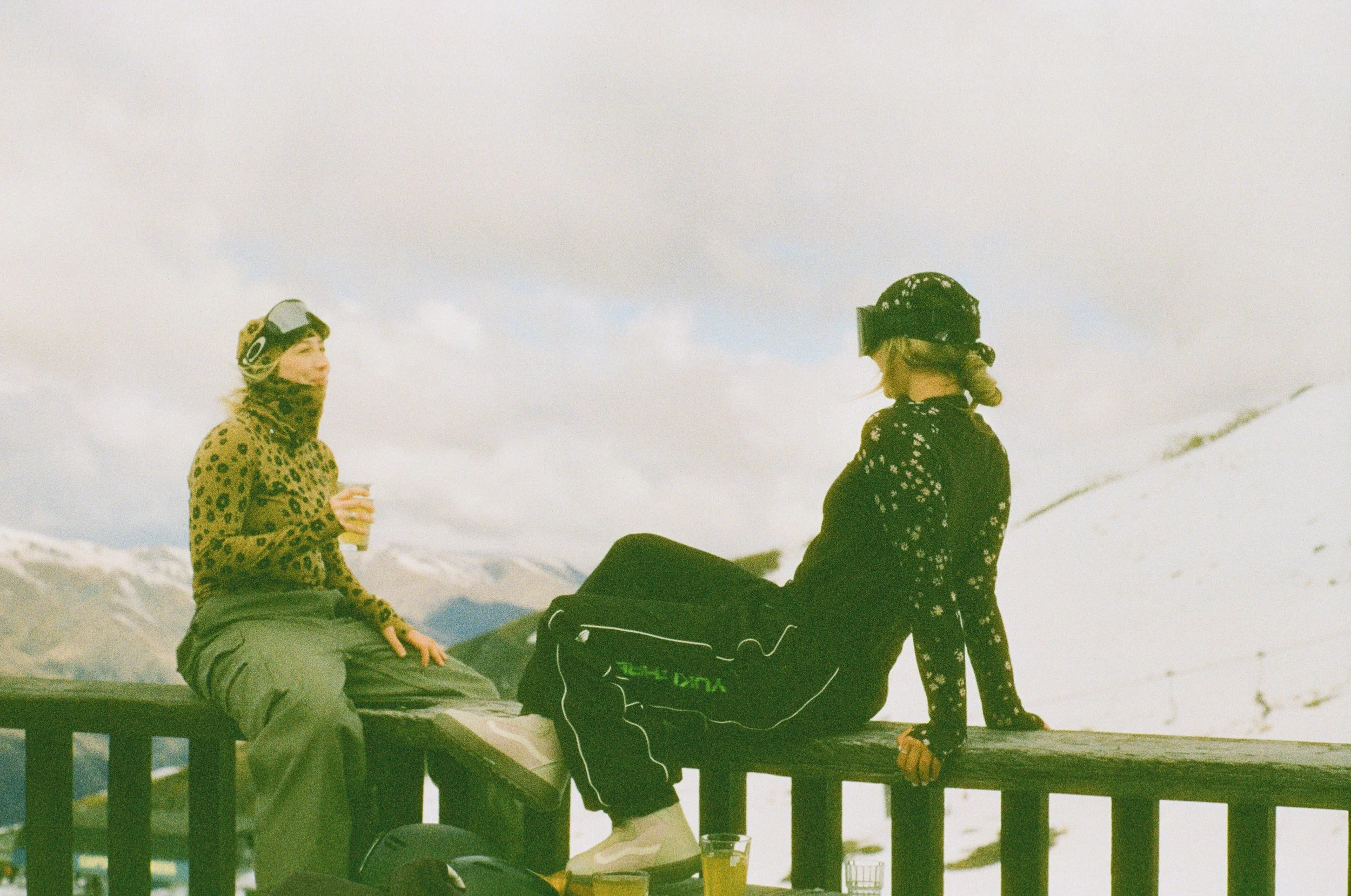 Two women sitting on a wooden railing outdoors in a snowy mountainous area, wearing winter clothing and ski helmets, with glasses of beer in front of them.