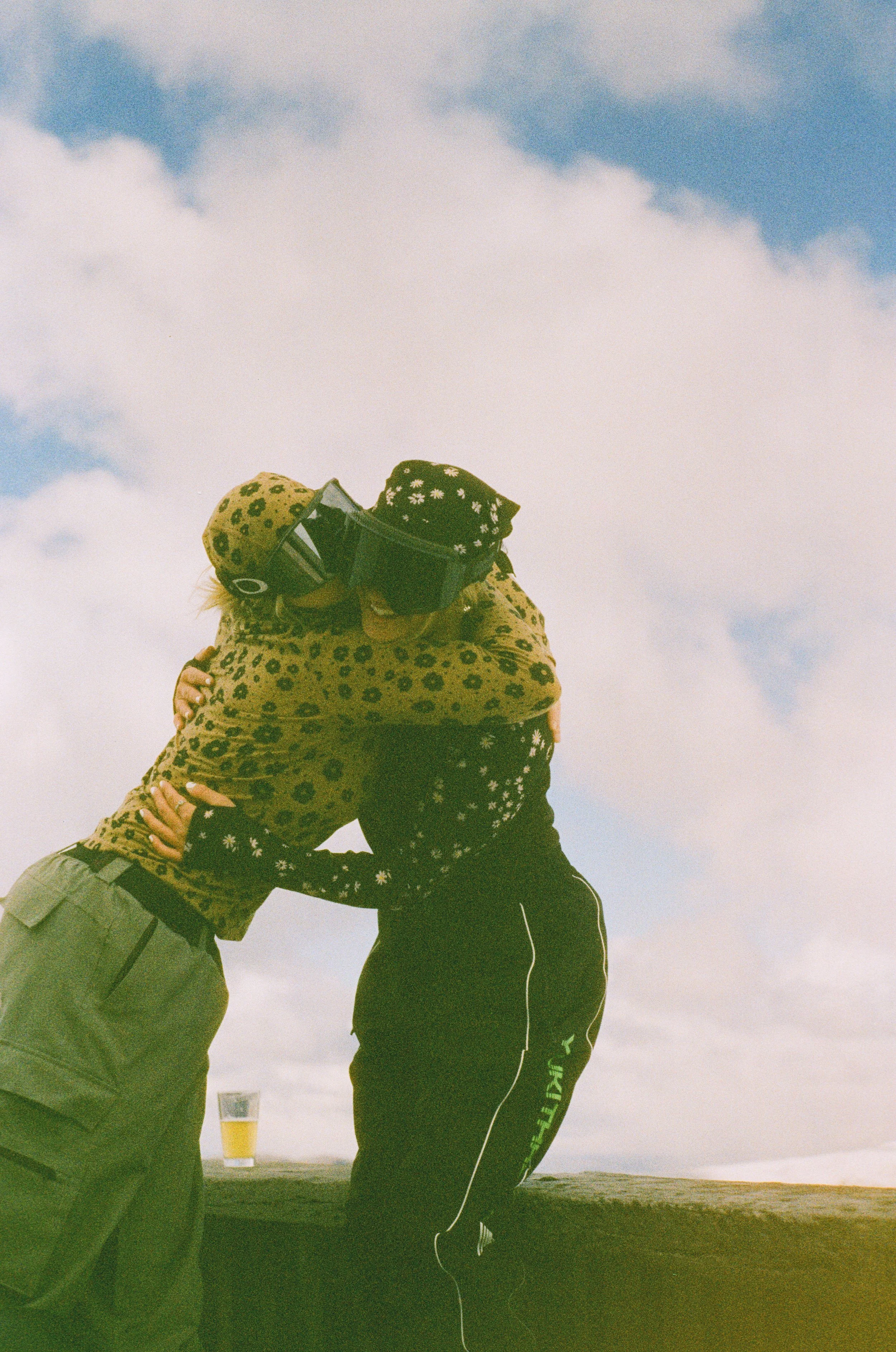 Two people hugging in front of a cloudy sky, with a drink on a table in the background.