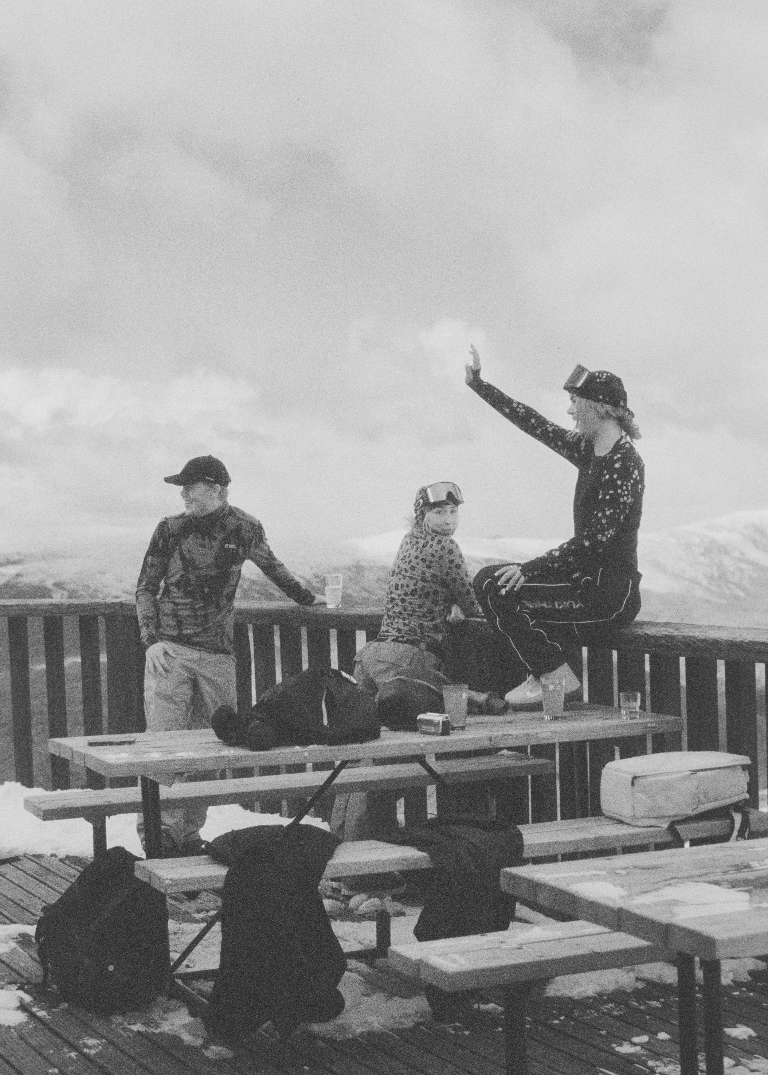 Group of people sitting and talking outdoors on a wooden deck, some standing, with snow and cloudy sky in the background.