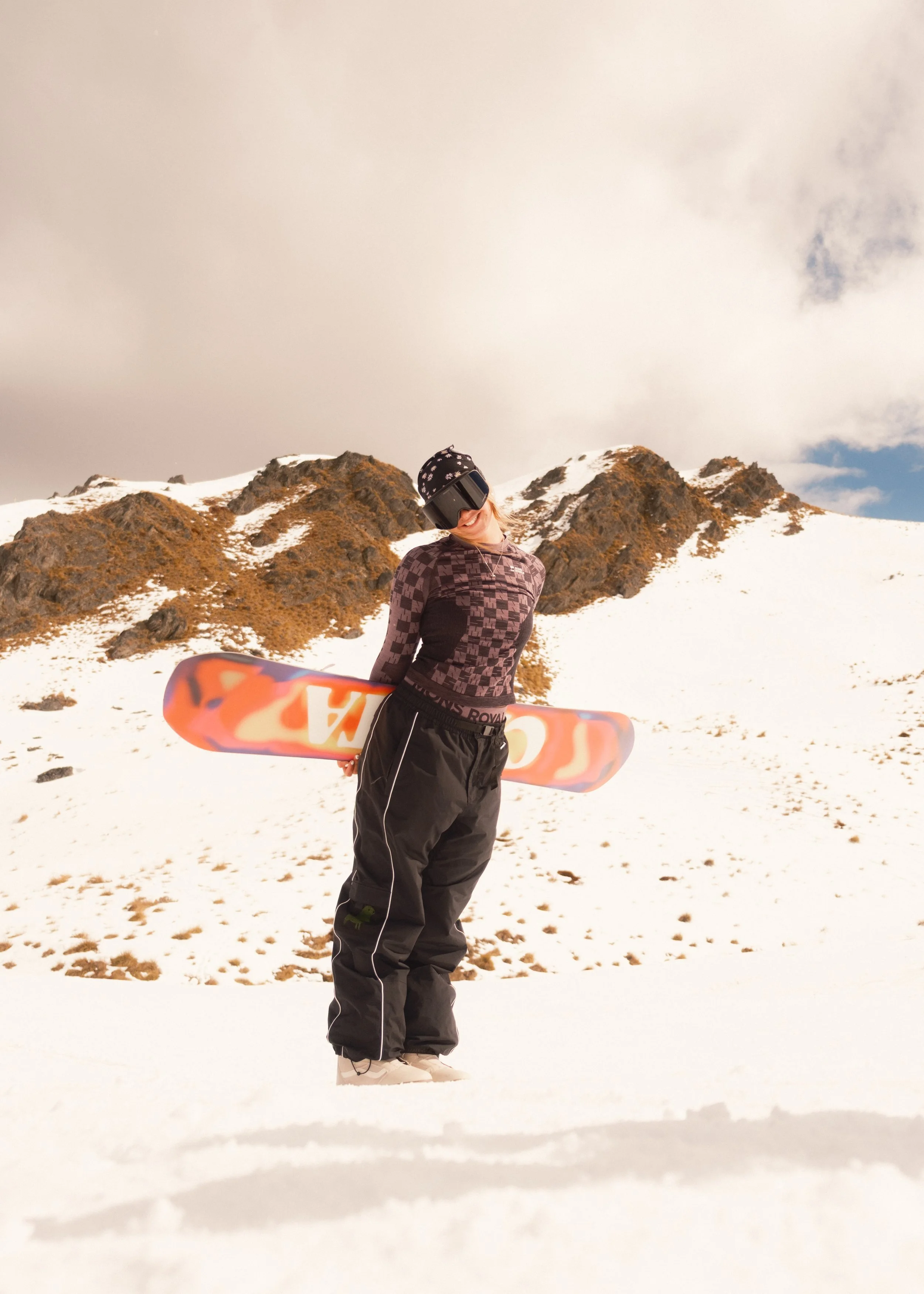 Person standing in snow-covered mountain landscape holding a snowboard, wearing a helmet, goggles, and winter athletic clothing.
