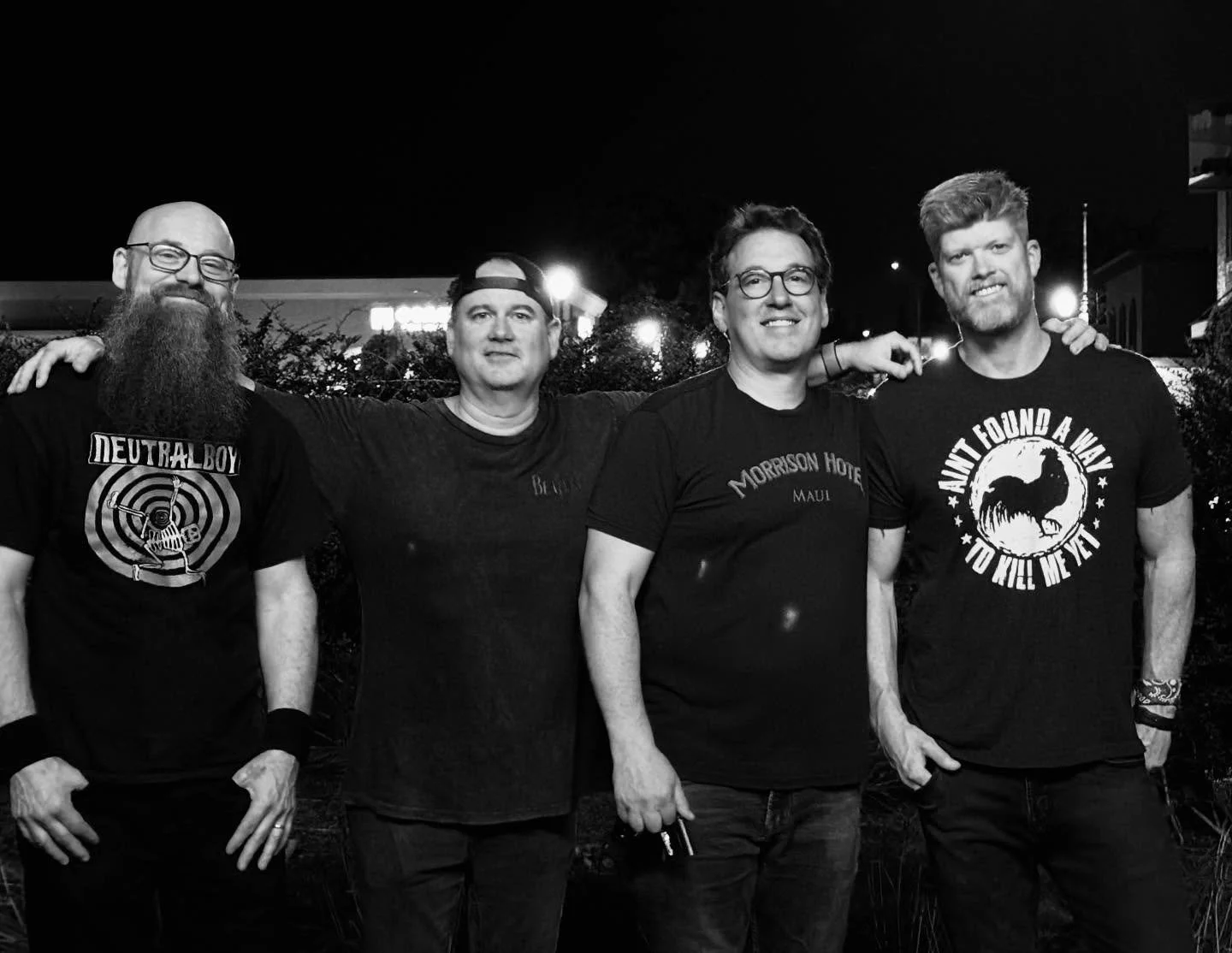 Four men standing outdoors at night, with their arms around each other, smiling at the camera. They are wearing casual t-shirts.