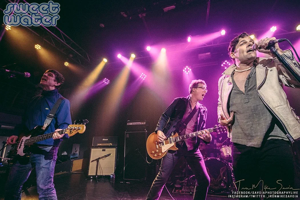 A band performing live on stage with purple and yellow stage lights, featuring a singer with a microphone and two guitarists, one playing bass and the other playing an electric guitar.