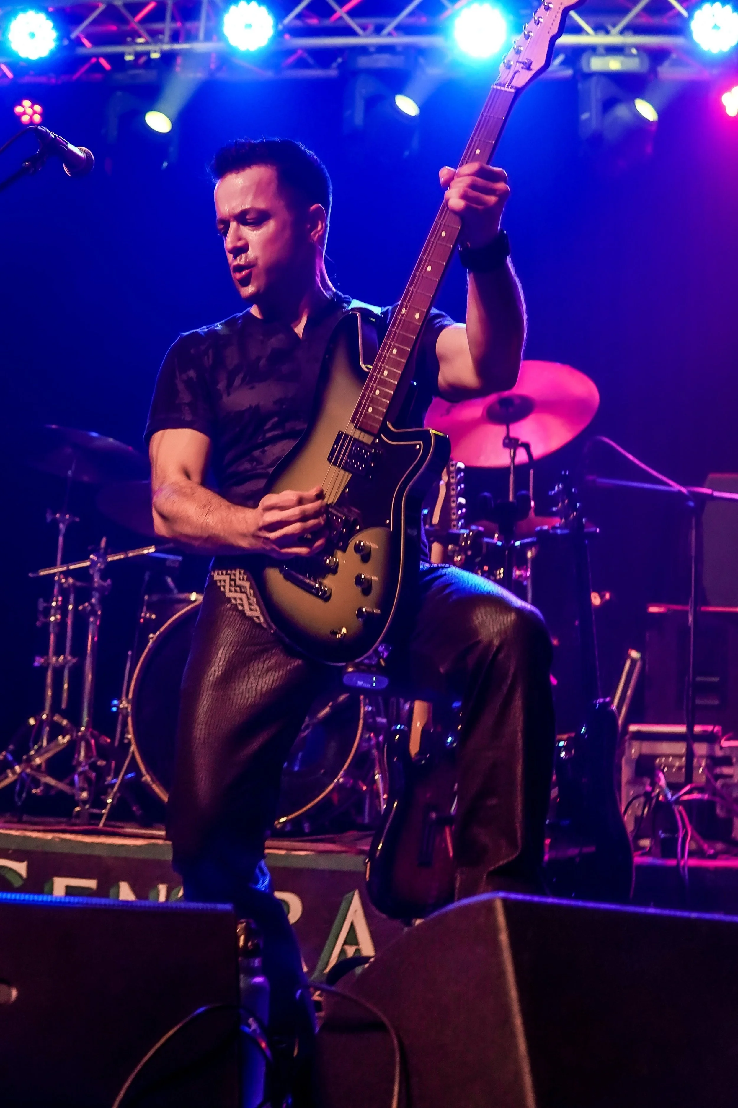 A man is playing an electric guitar on stage during a live music performance with colorful stage lighting, drums in the background, and a microphone near him.