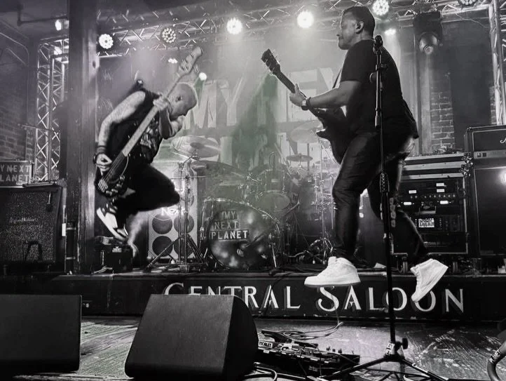 Two musicians playing guitars on stage at a concert, with drums in the background and stage lights overhead, in black and white.