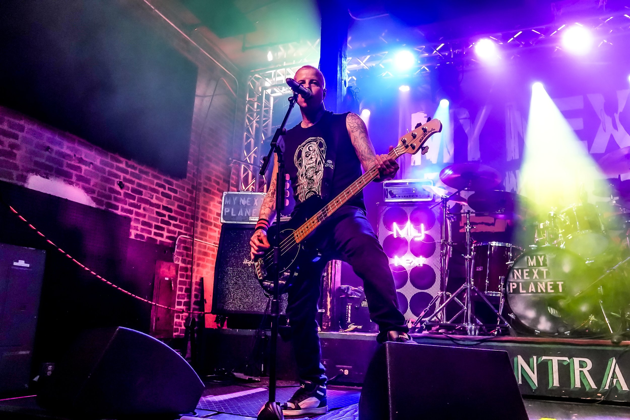 Musician performing on stage with a bass guitar, colorful stage lighting, and a brick wall background, with a neon sign reading 'MY NEXT PLANET'.
