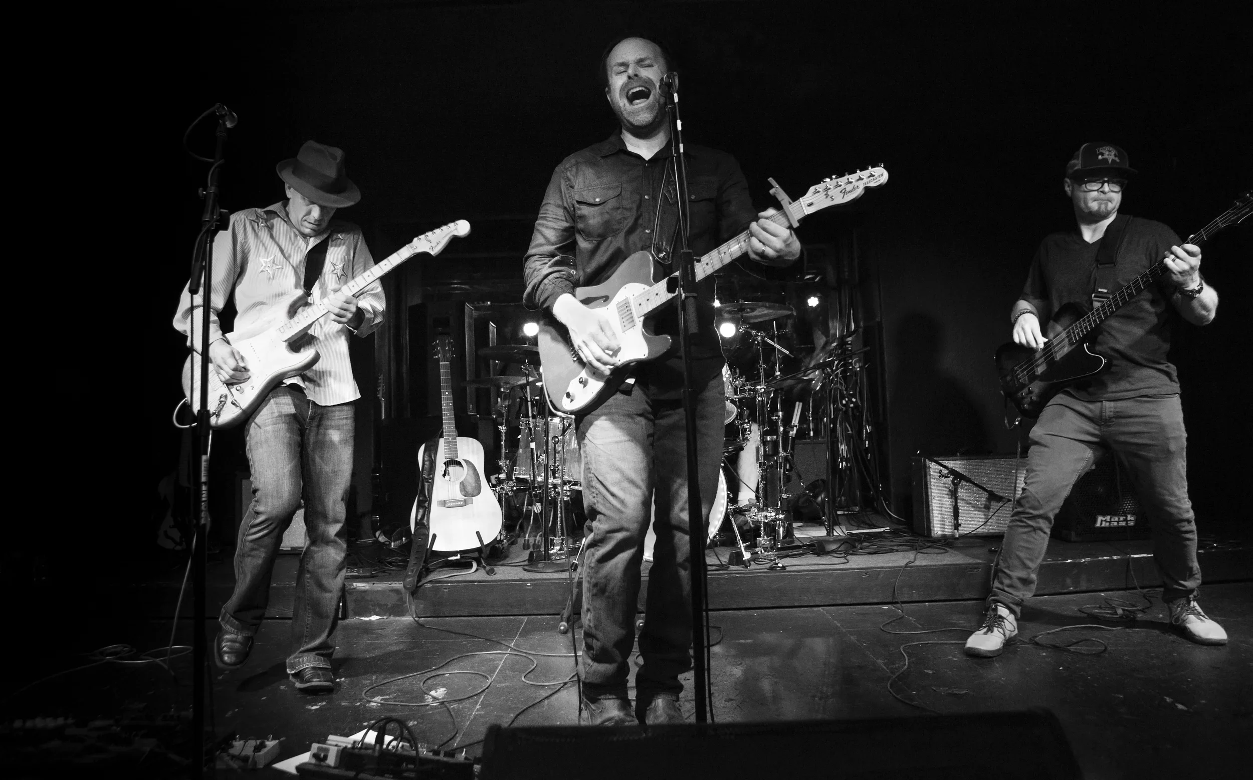 A black and white photograph of a band performing on stage. The lead singer is playing an electric guitar and singing passionately with his eyes closed. Two other band members are playing guitars beside him, and a drum set is visible in the backgroun