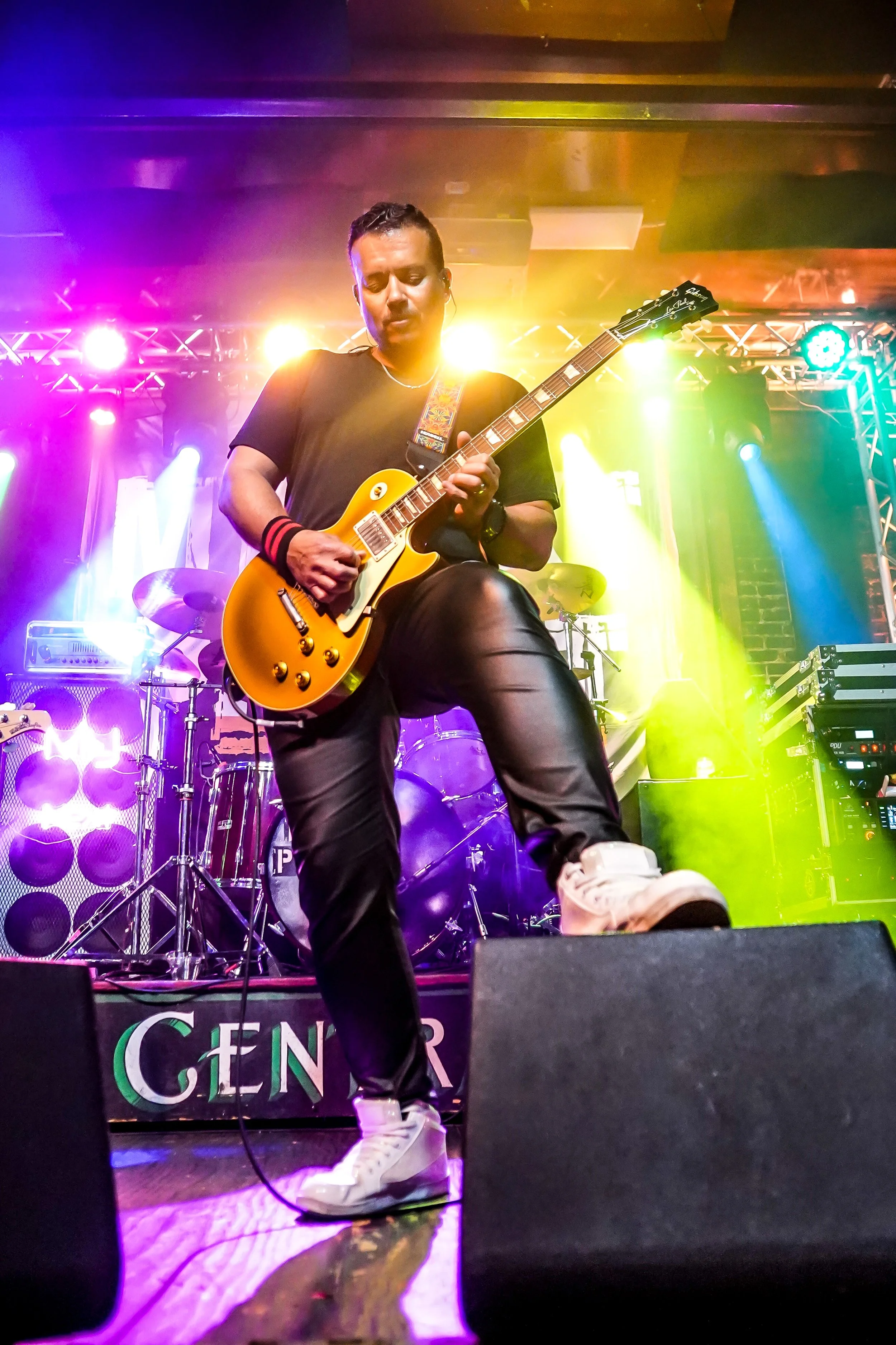 A male guitarist performing on stage with colorful stage lighting, wearing a black t-shirt, black pants, and white sneakers, with a drum set in the background.
