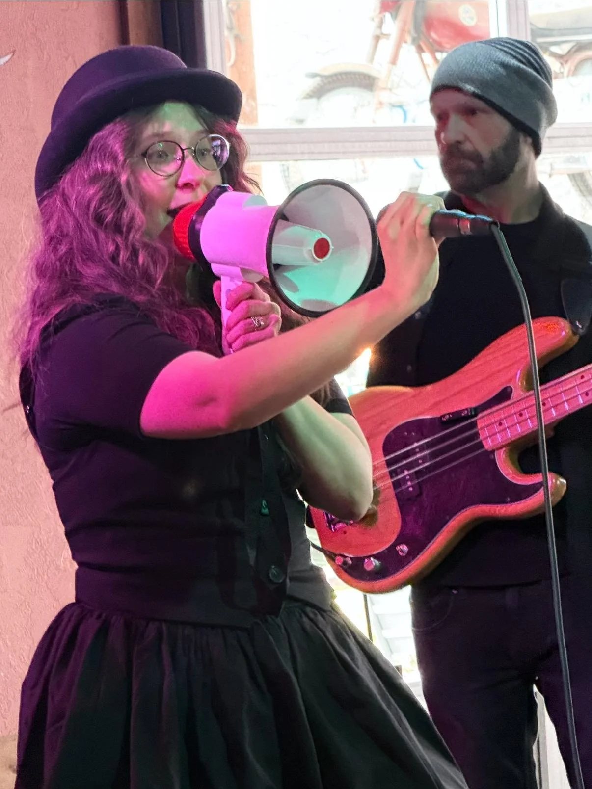 A woman with long curly hair, glasses, and a black hat holding a megaphone, with a man playing a bass guitar beside her. She is wearing a black dress with a full skirt. The man is wearing a gray beanie and a dark jacket. There is a window in the back