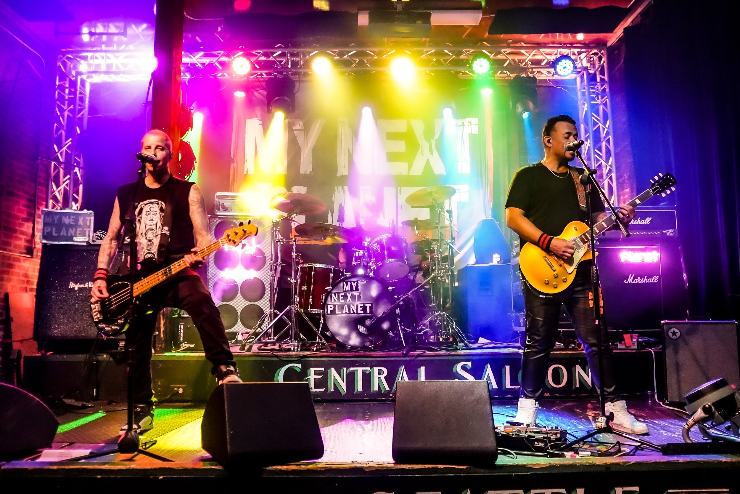 A band performs live on stage with colorful stage lighting. Two guitarists sing into microphones, with one on the left and the other on the right. The drummer is in the background, and the stage is set with amplifiers and musical equipment. The backd