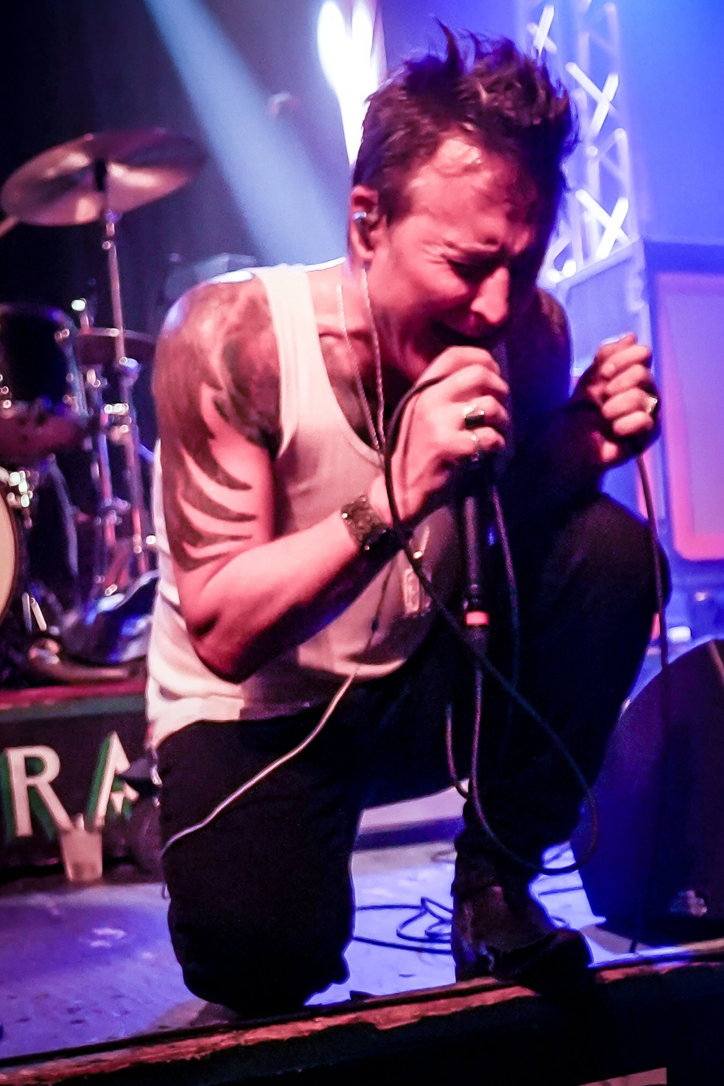 A male singer with tattoos, wearing a sleeveless shirt, passionately performing into a microphone on stage with colorful lights and a drum set in the background.