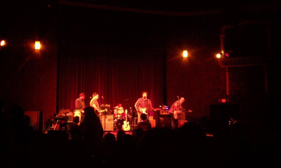 Band performing live on stage at a dimly lit concert venue, with audience silhouettes in the foreground.