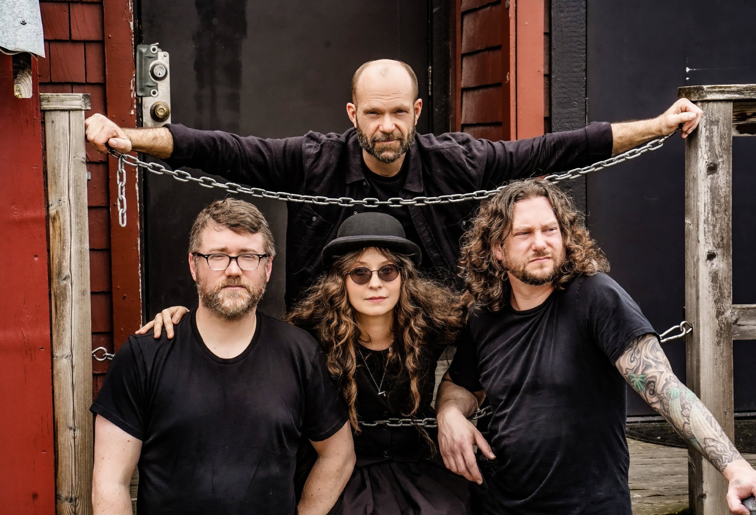 Group of four people, three men and one woman, standing outside in front of a black door and red wooden siding. The man in the back has a bald head, beard, and is wearing a black jacket. The woman has curly brown hair, sunglasses, and a hat. The man on the left has glasses, a beard, and a black shirt. The man on the right has curly hair, a beard, and tattoos on his arm.