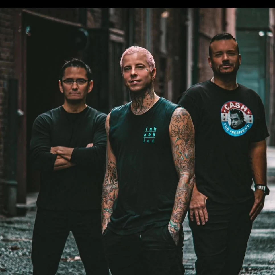 Three men standing in an alleyway with brick walls. The man in the middle has pink hair, tattoos on his arms, and wears a sleeveless black shirt. The man on the left has glasses, short dark hair, and arms crossed. The man on the right has short dark 
