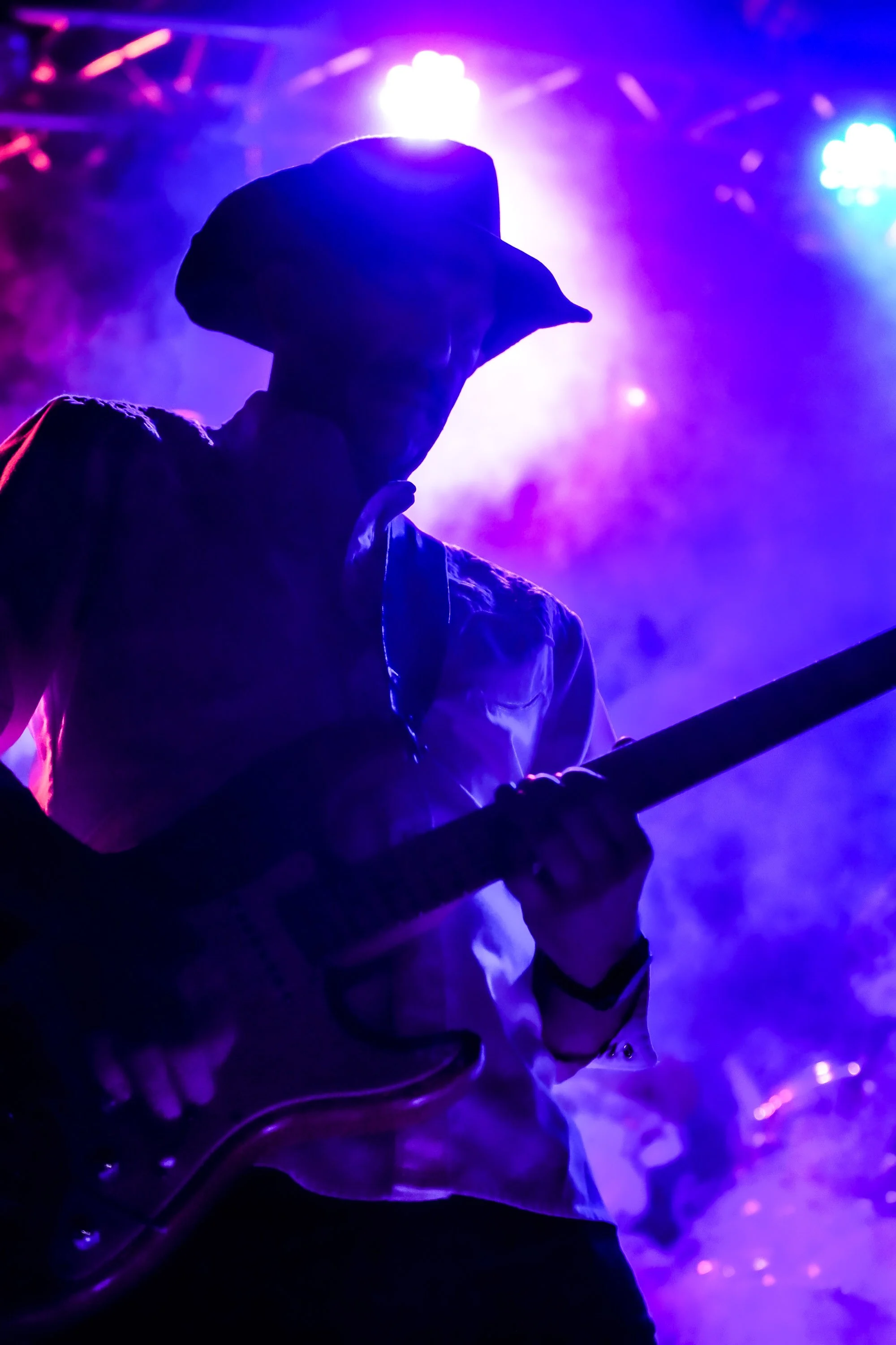 Silhouetted person in a hat, holding a guitar, surrounded by colorful stage lighting and smoke effects at a concert or performance.