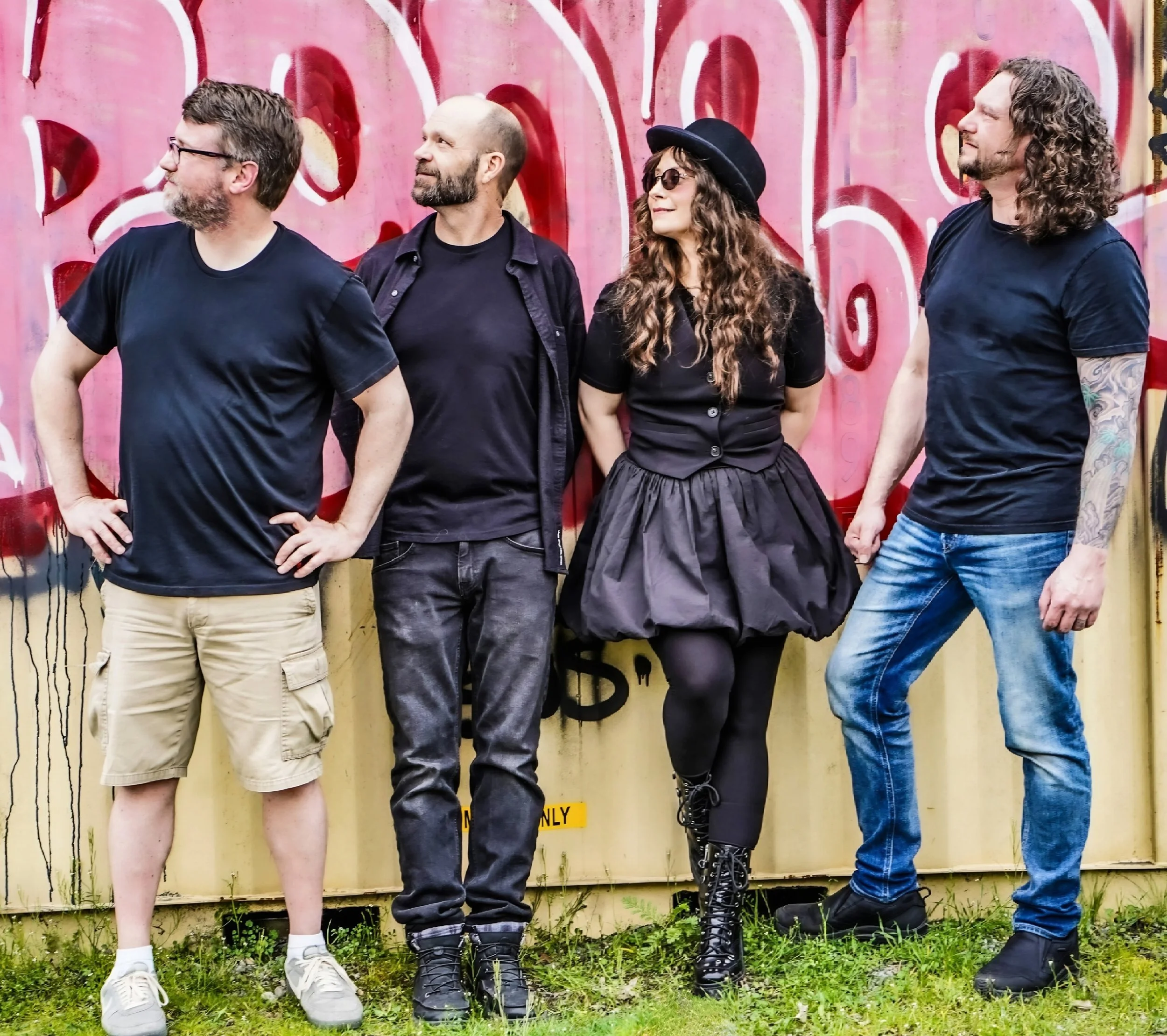 Four people standing side by side outdoors in front of a graffiti wall. The first person on the left is a man in a black t-shirt and khaki shorts, the second is a bearded man in black with a jacket, the third is a woman with curly hair wearing a blac