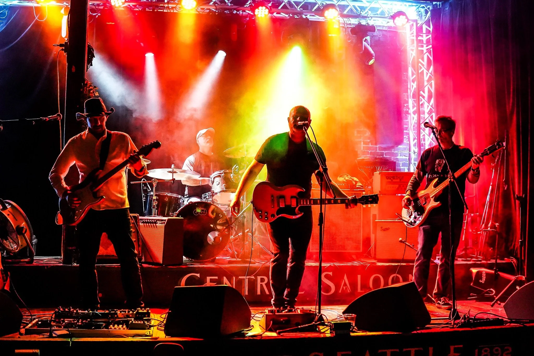 A band performing on stage with colorful lighting, including at least four musicians playing guitars and drums, with a brick wall and stage lighting in the background.