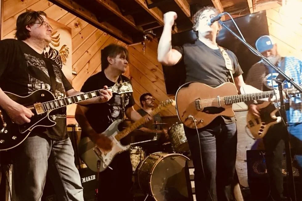 A band performing live on stage, with four members playing guitars and drums in a wooden-paneled venue.