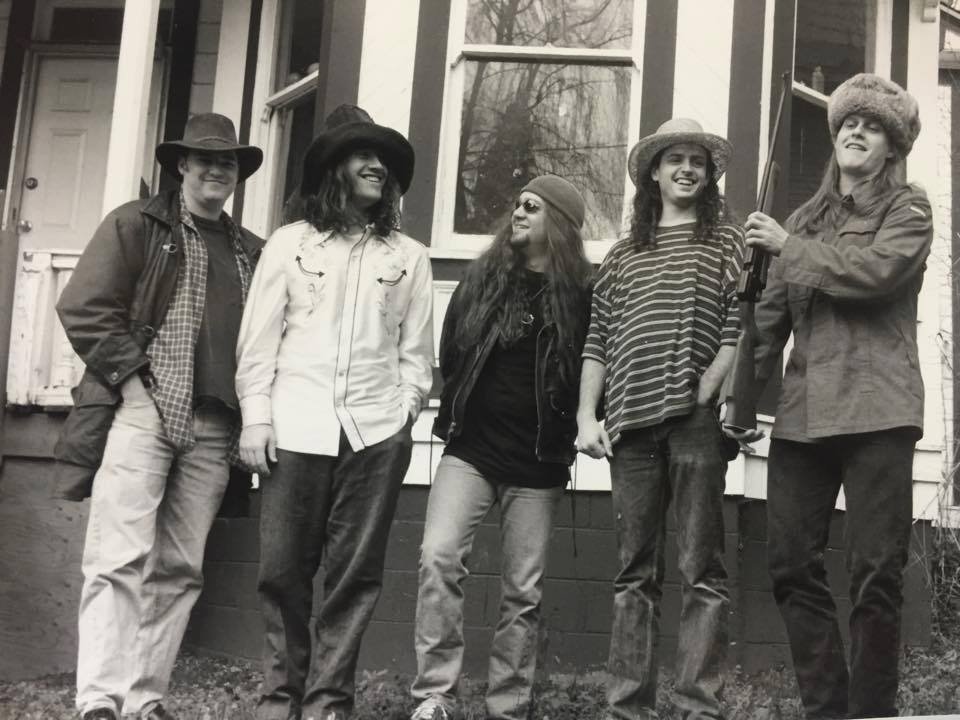 Group of five young people standing outside a house, smiling, with two of them holding a rifle.
