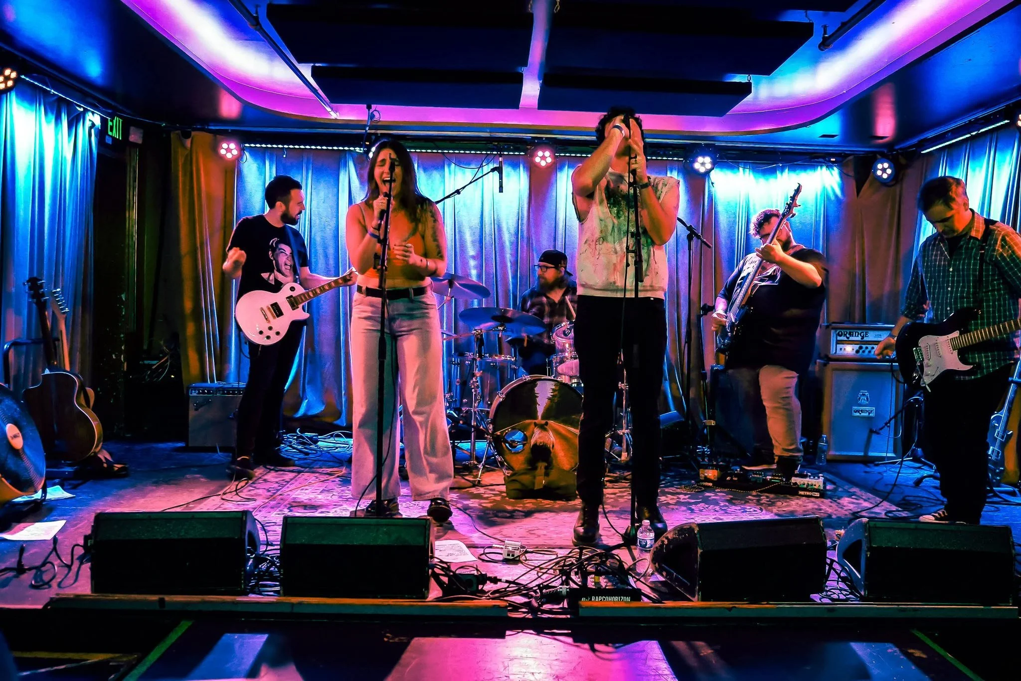 Band performing on stage with colorful lighting, guitars, and microphones, with a curtain backdrop.