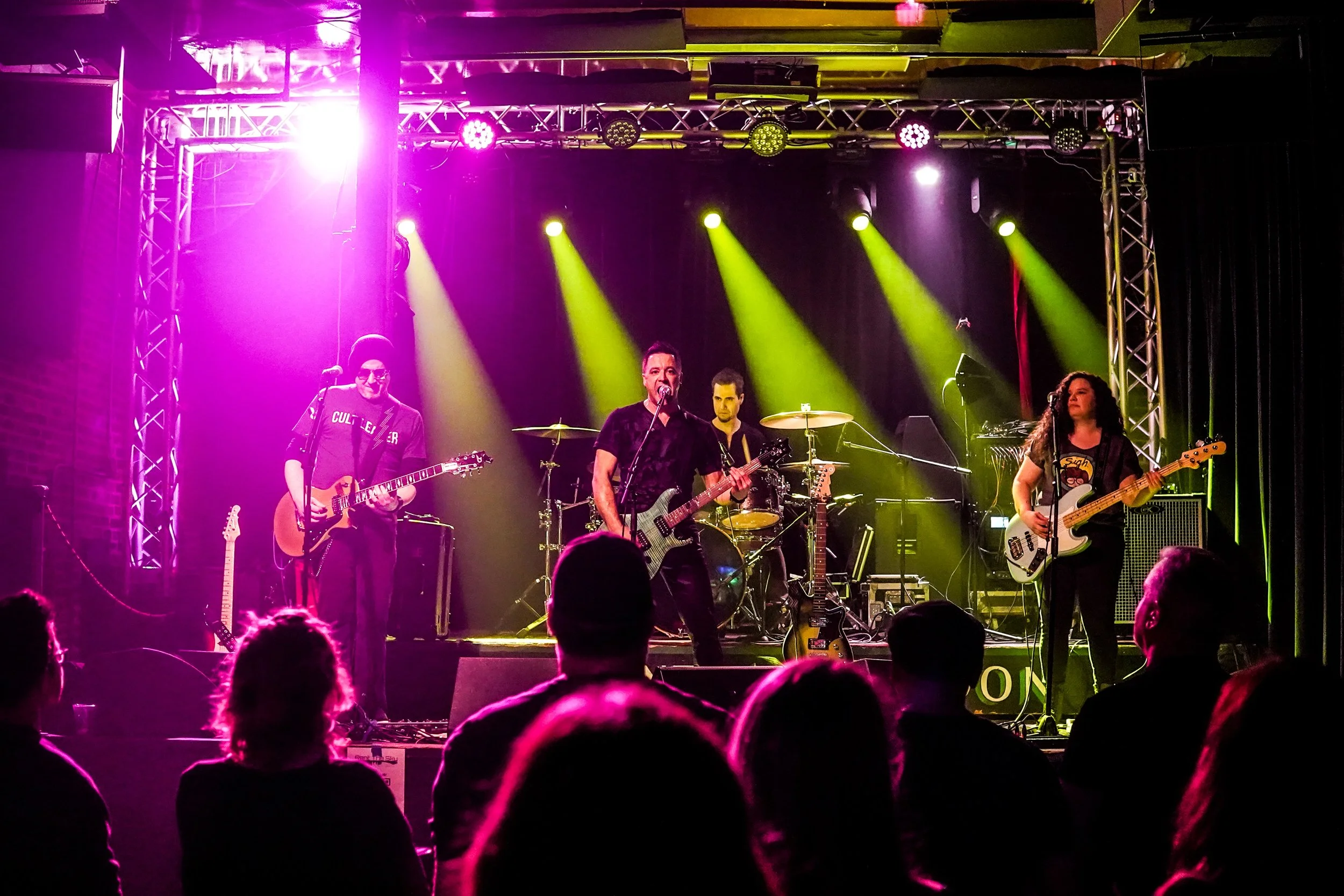 A musical band performing on stage with colorful lights, including purple and yellow, illuminating the scene. The band members are playing guitars and drums, and an audience is visible in the foreground.