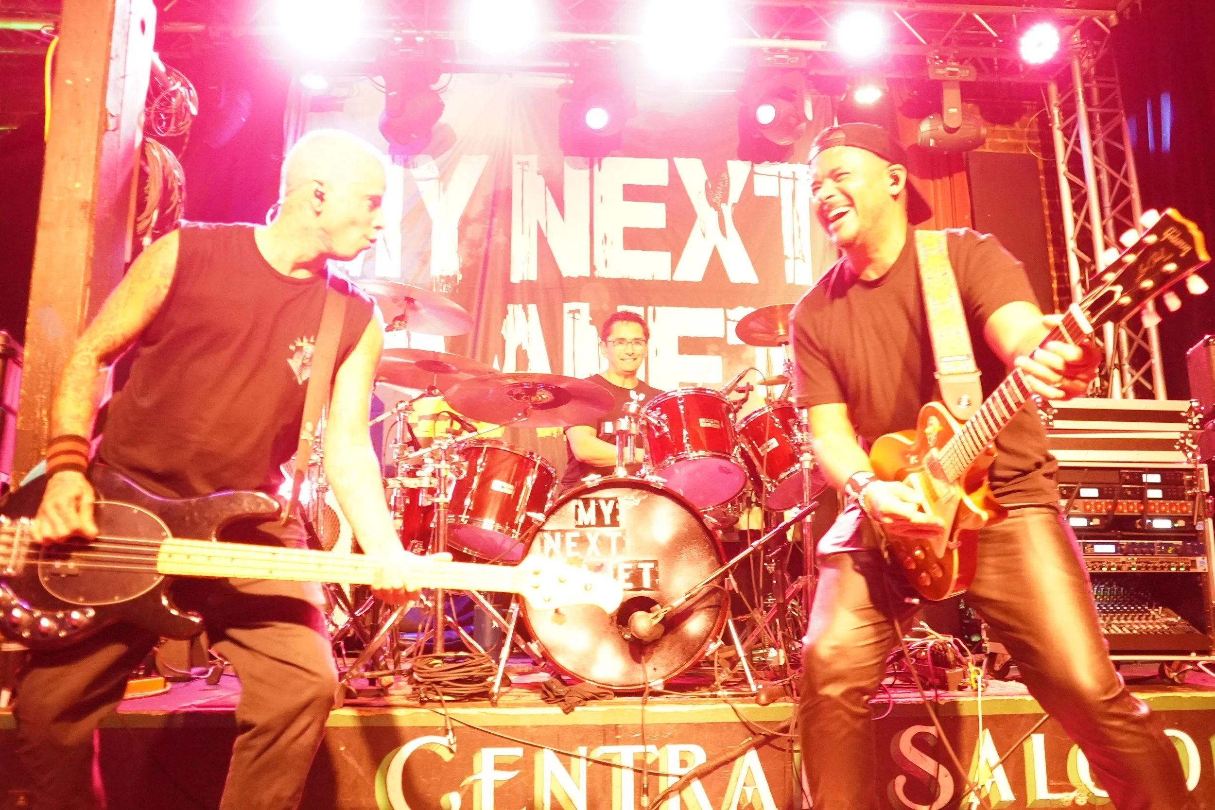 Three musicians performing on stage with bright lights, two guitar players and a drummer, with a backdrop that reads 'My Next Exit'.