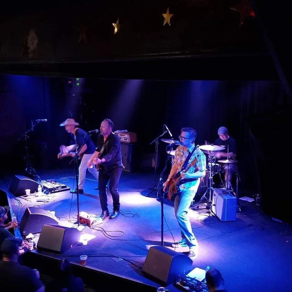 A band performs on stage with four members: a singer with a microphone, a guitarist with a hat, a bassist, and a drummer, under purple stage lighting, in a venue decorated with star-shaped ornaments hanging from the ceiling.