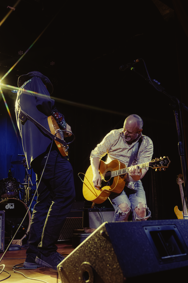 Two musicians performing on stage with guitars, one in a hat and the other with a shaved head, under stage lights.