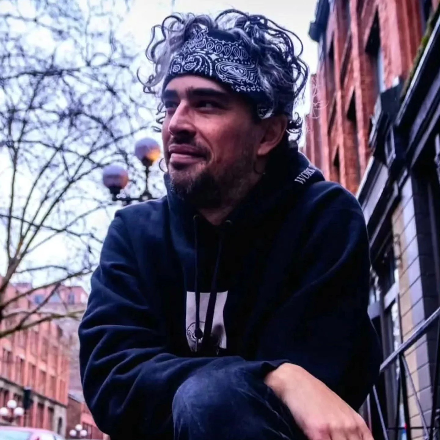 A man with curly hair, wearing a bandana and a black hoodie, sitting outdoors on a city street with brick buildings and leafless trees in the background.