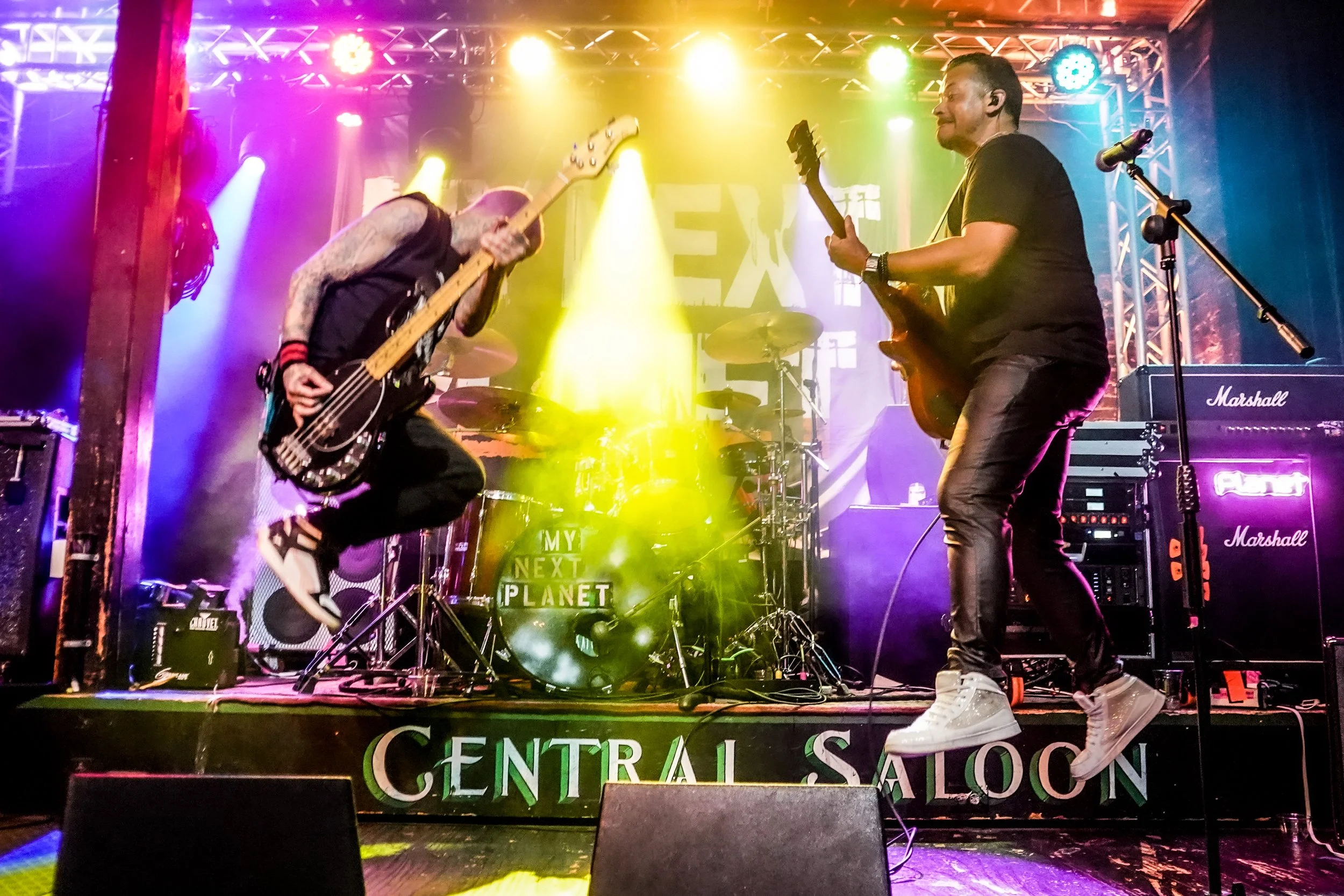 Two musicians performing on a stage with colorful stage lighting. One is playing bass guitar, the other playing an electric guitar. A drum set is visible in the background. The stage has a sign that reads "Central Saloon".