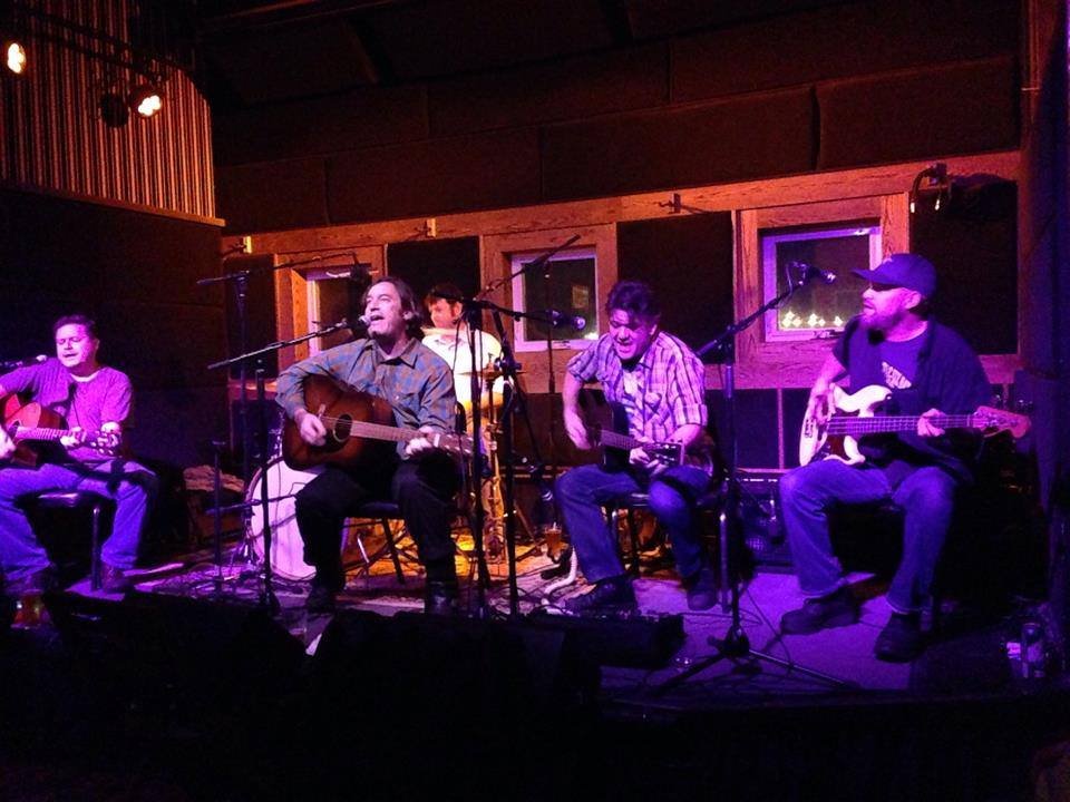 Five musicians performing on a stage in a dimly lit venue. Three are playing guitars, and one is singing into a microphone. They are seated close together with a wooden backdrop and purple lighting.