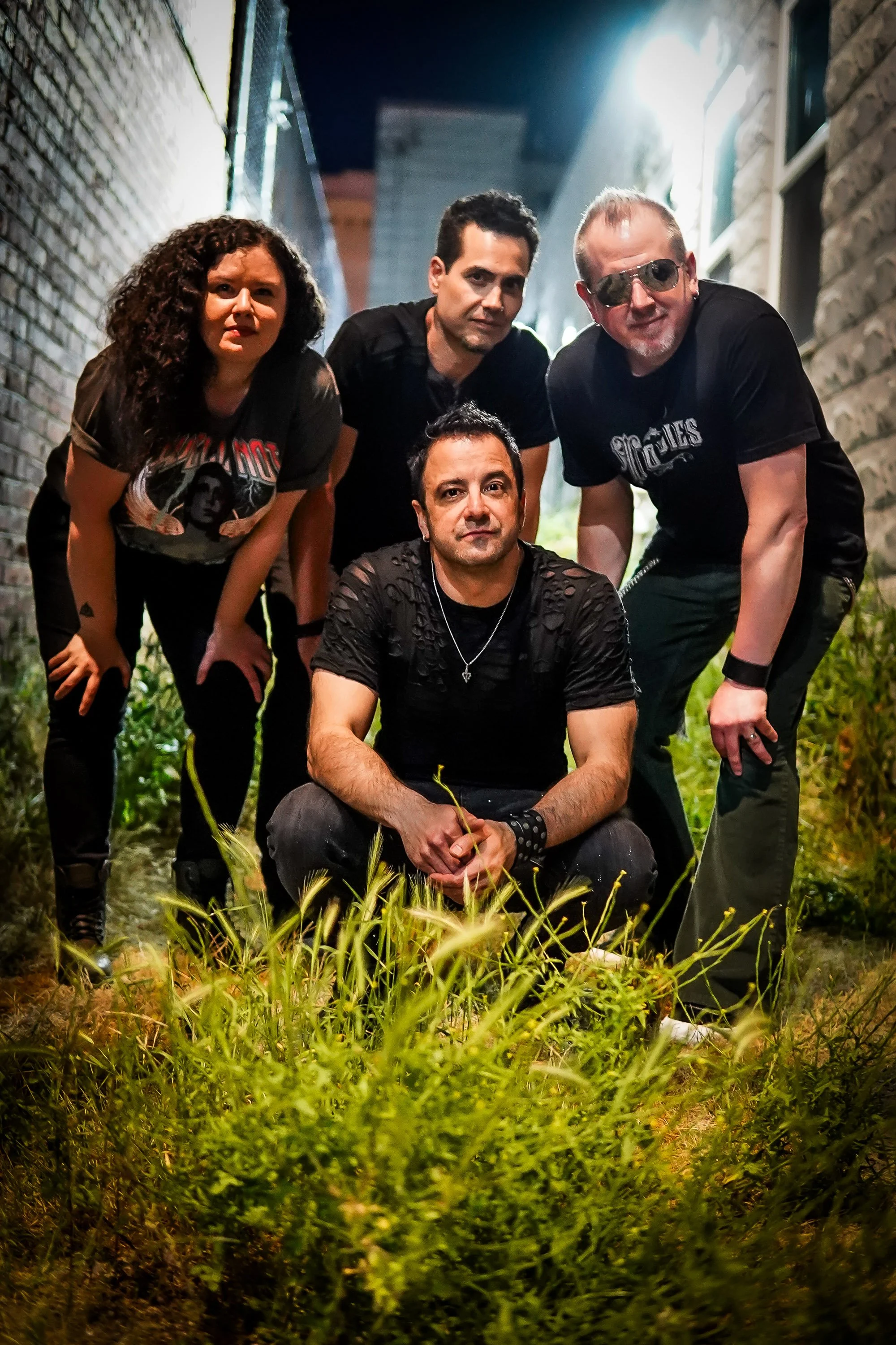 Group of five people posing in a narrow alleyway with brick walls on either side. They are crouching or kneeling, and are surrounded by green grass and plants on the ground. The person in front is seated, wearing a black shirt with ripped sleeves, and has their hands clasped in front of them. The other four are standing or leaning behind, with diverse expressions and styles, including sunglasses and band t-shirts. The lighting is moody with some bright light coming from the top right.