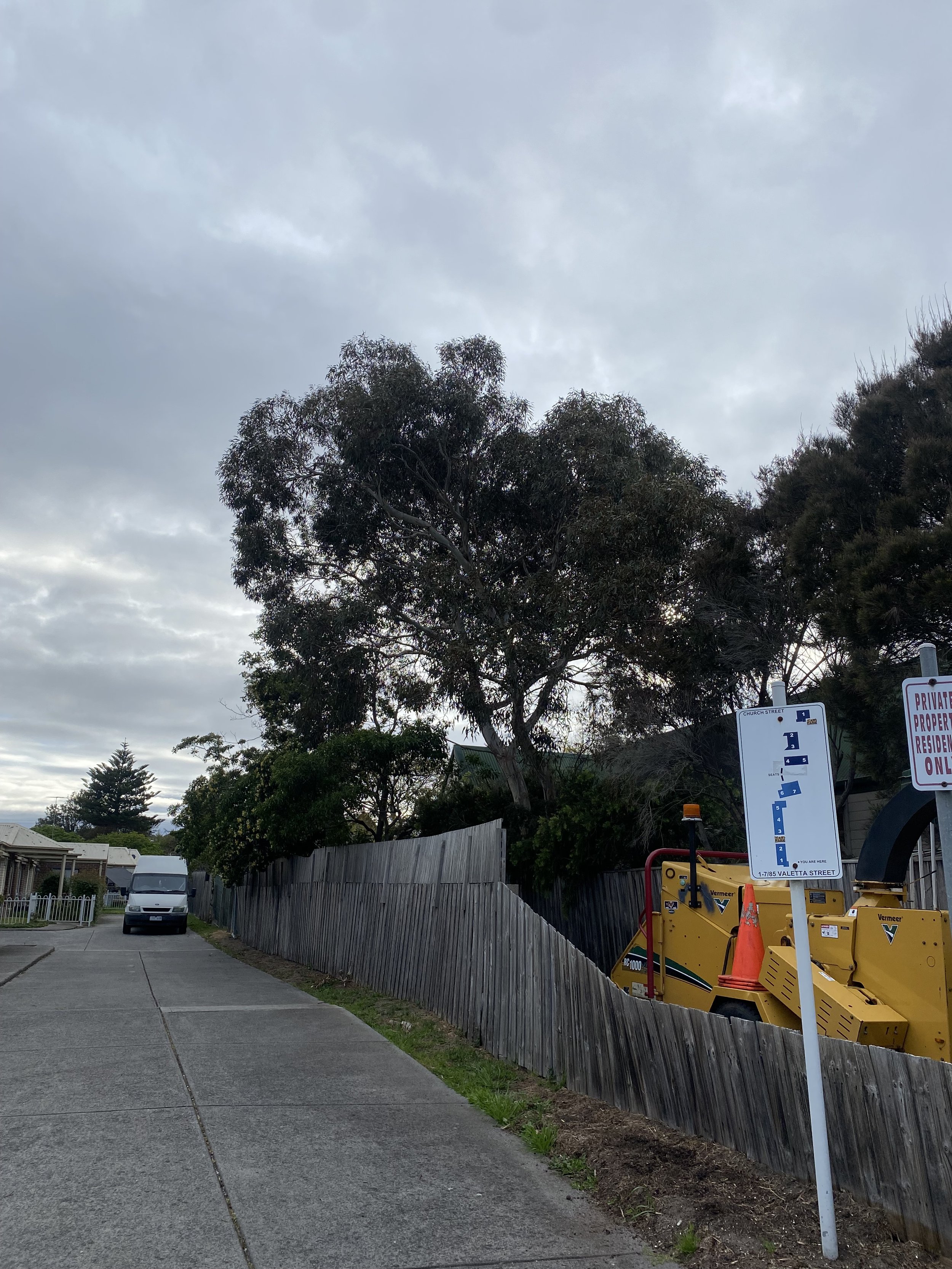 A sidewalk with a parked van in front of a wooden fence, trees, and a cloudy sky. There are street signs and a construction vehicle nearby.