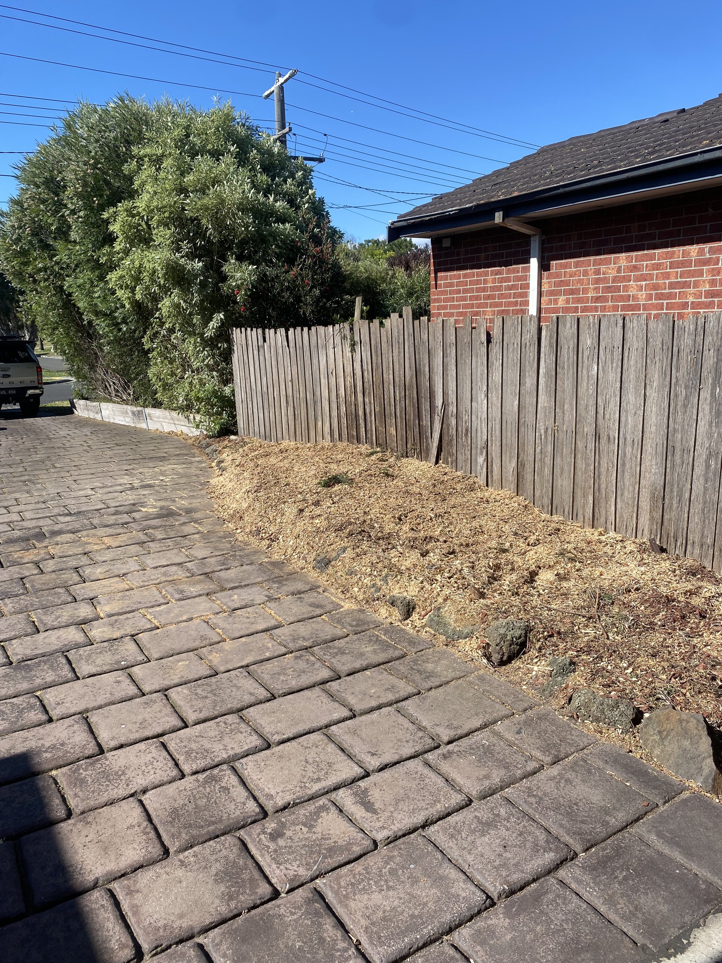 A cobblestone sidewalk beside a wooden fence, with a brick house on the right side and a large green bush behind the fence; clear sky and power lines overhead.