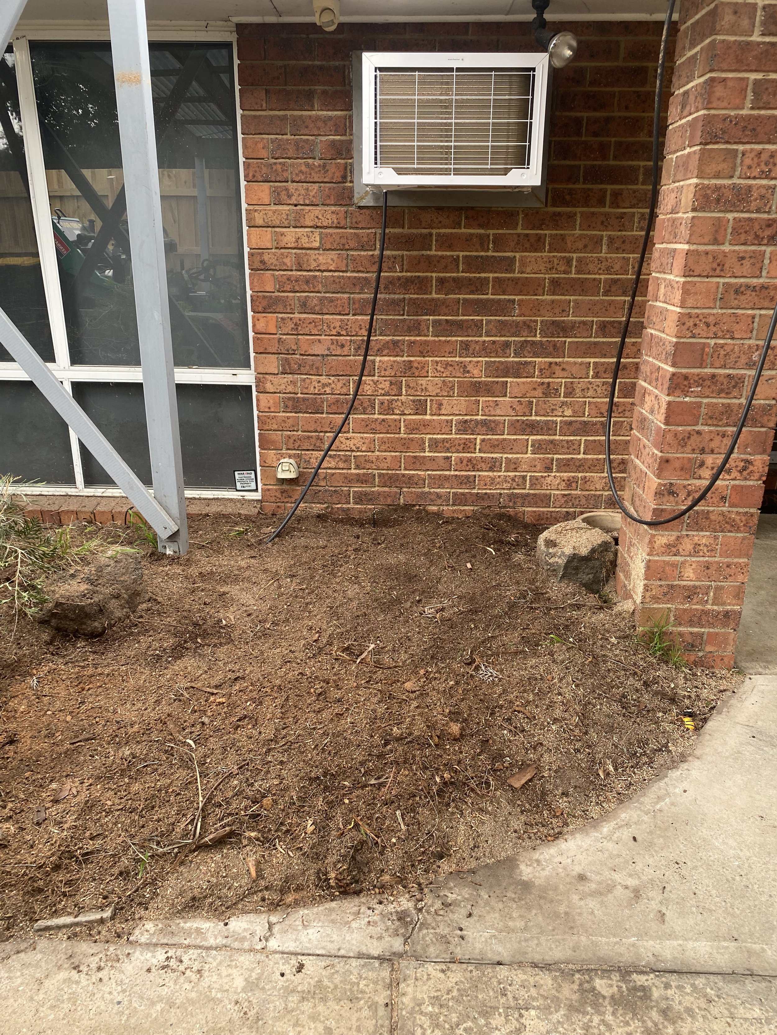 A backyard area with a dirt patch next to a concrete sidewalk with a brick house wall and a window air conditioning unit, wires hanging down, and some rocks and plants.