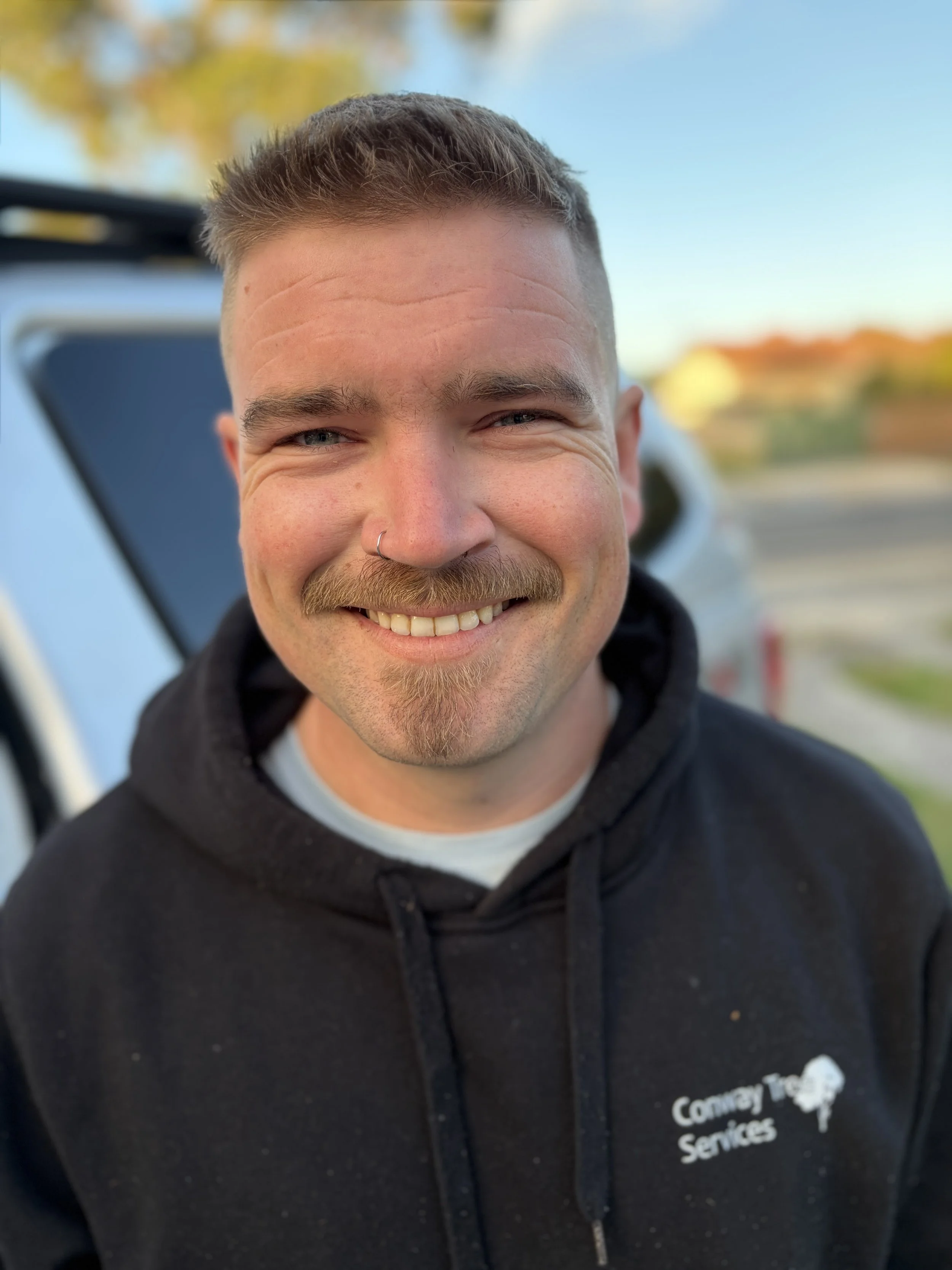 Close-up of a smiling man with short hair, facial hair, nose piercing, wearing a black hoodie with 'Conway' and 'Services' printed on it, standing outdoors with blurred background.