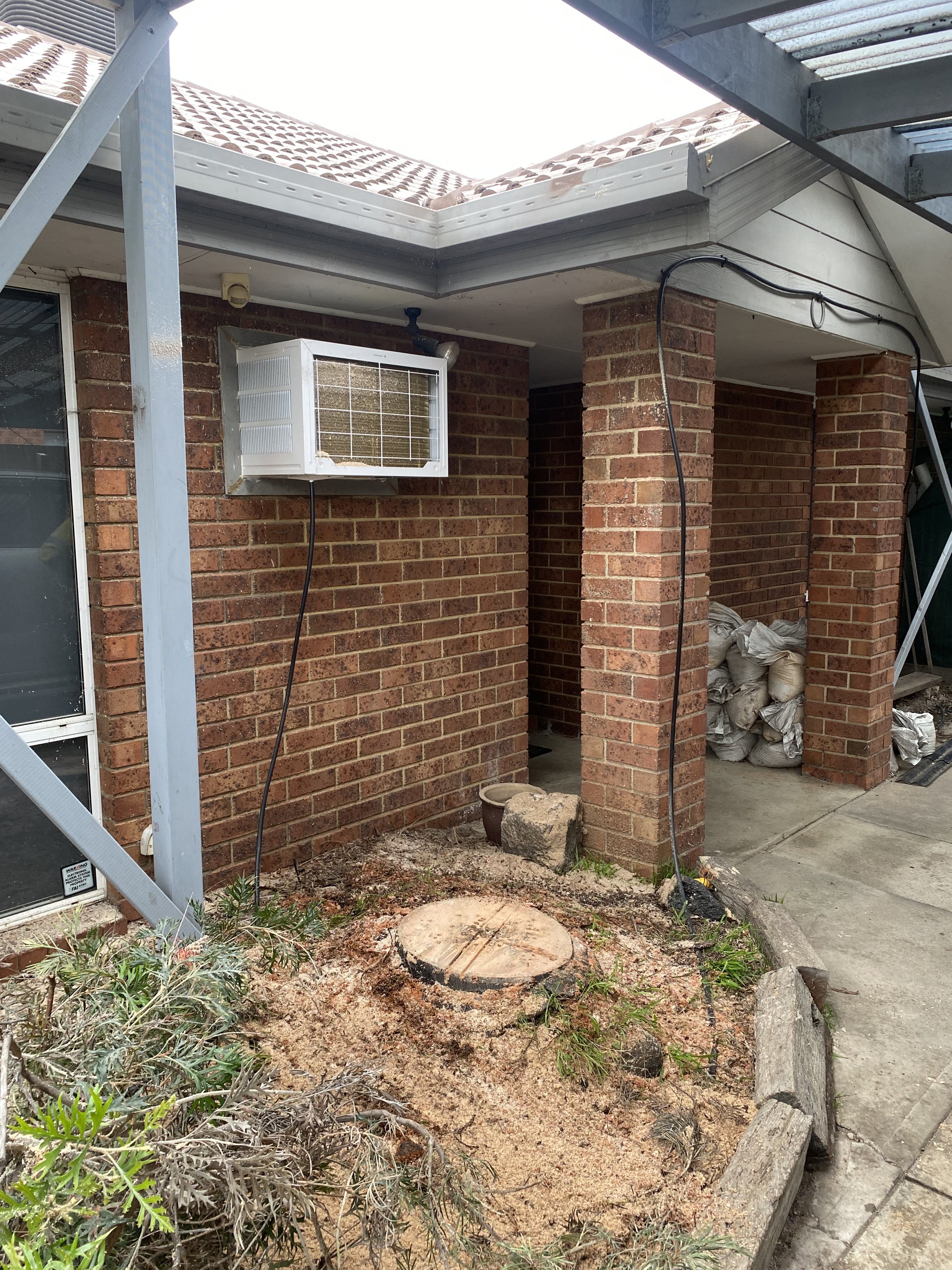 Exterior view of a brick house with a white window air conditioning unit installed in the wall. There is construction or landscaping work ongoing, with a large tree stump, dirt, plants, and piles of sandbags on the ground. Metal scaffolding is presen